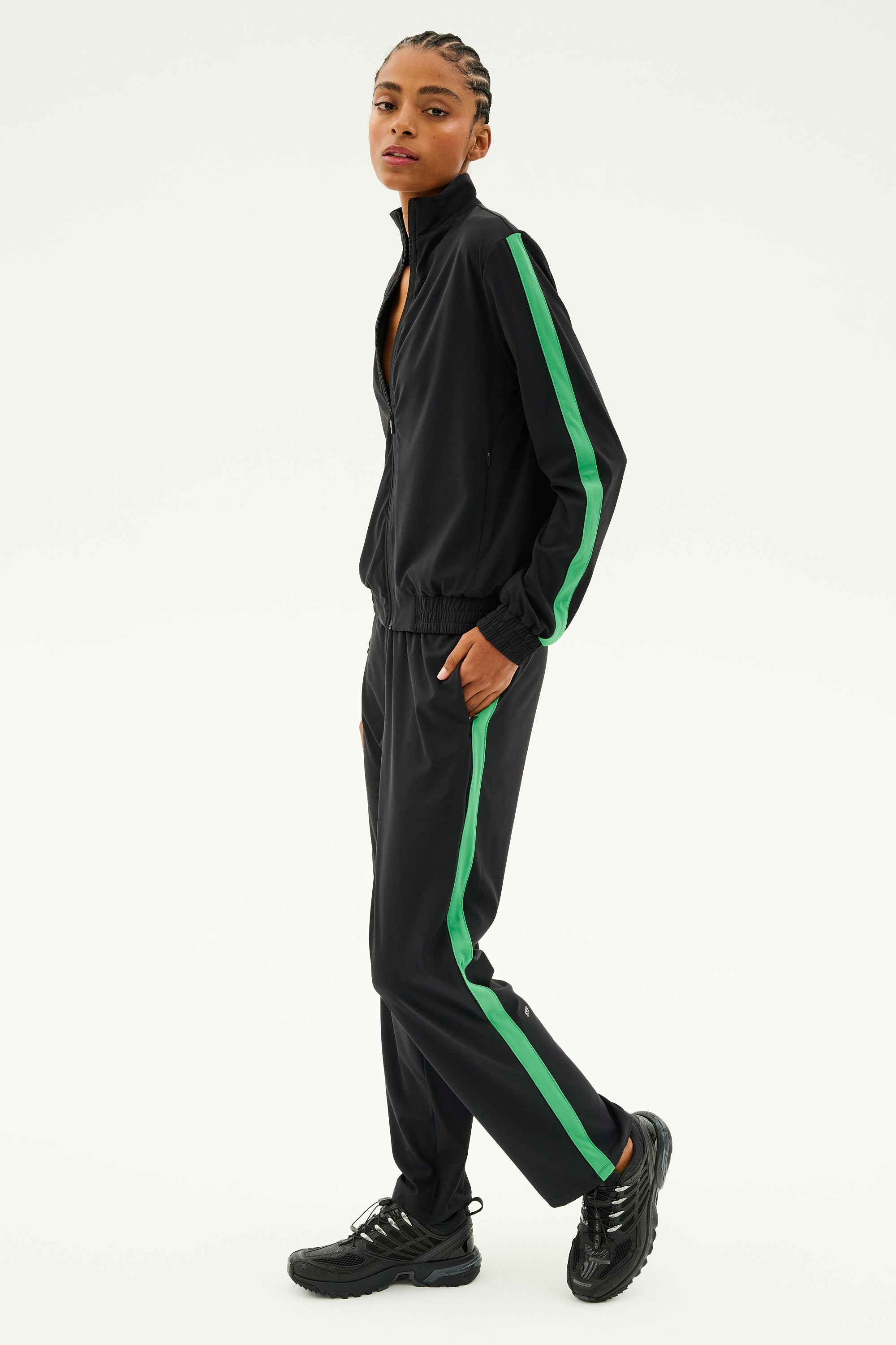 A person wearing the SPLITS59 Max Rigor Track Jacket in Black/Grass, featuring green stripes and crafted from rigor fabric, stands with one hand in their pocket against a plain background. Black sneakers complete this stylish workout piece.