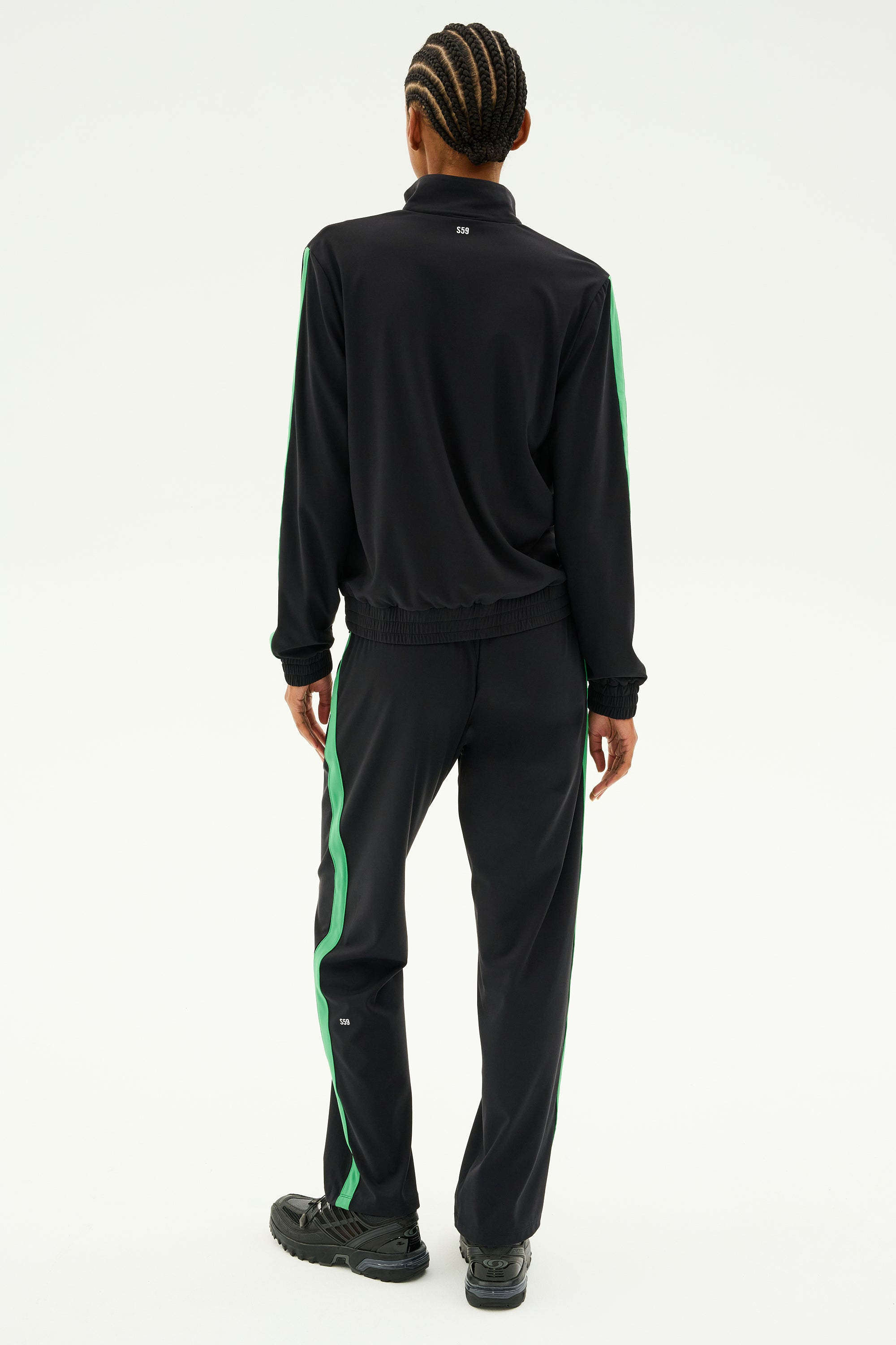 A person with braided hair is standing and facing away, dressed in the Max Rigor Track Jacket - Black/Grass by SPLITS59. This sleek tracksuit features green stripes, making the jacket the standout centerpiece of this chic workout ensemble that blends style and function seamlessly.