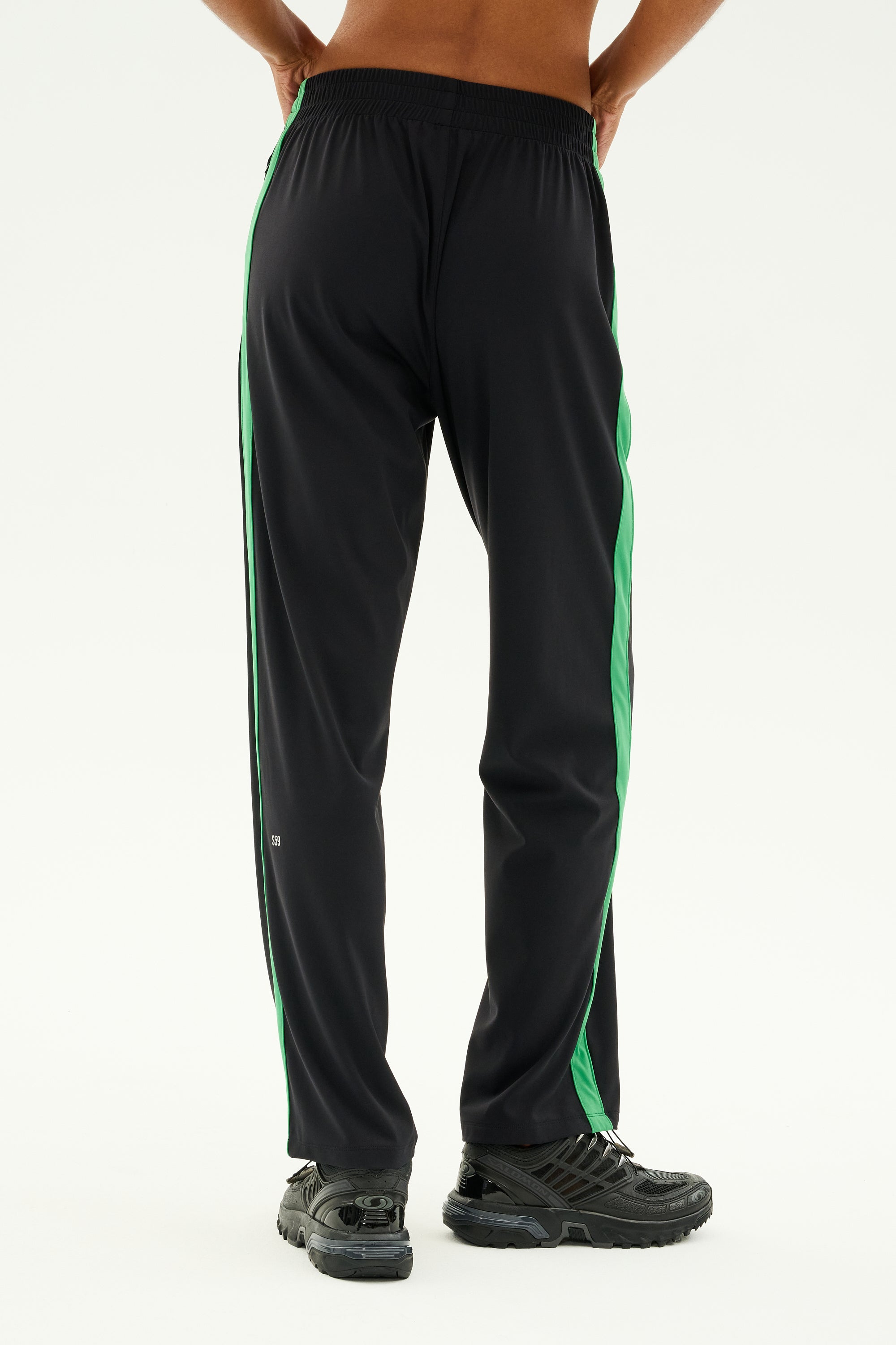 A person in the Max Rigor Track Pant - Black/Grass by SPLITS59 stands with their back turned and hands on hips, epitomizing the spirit of outdoor workouts in sleek black athletic shoes.