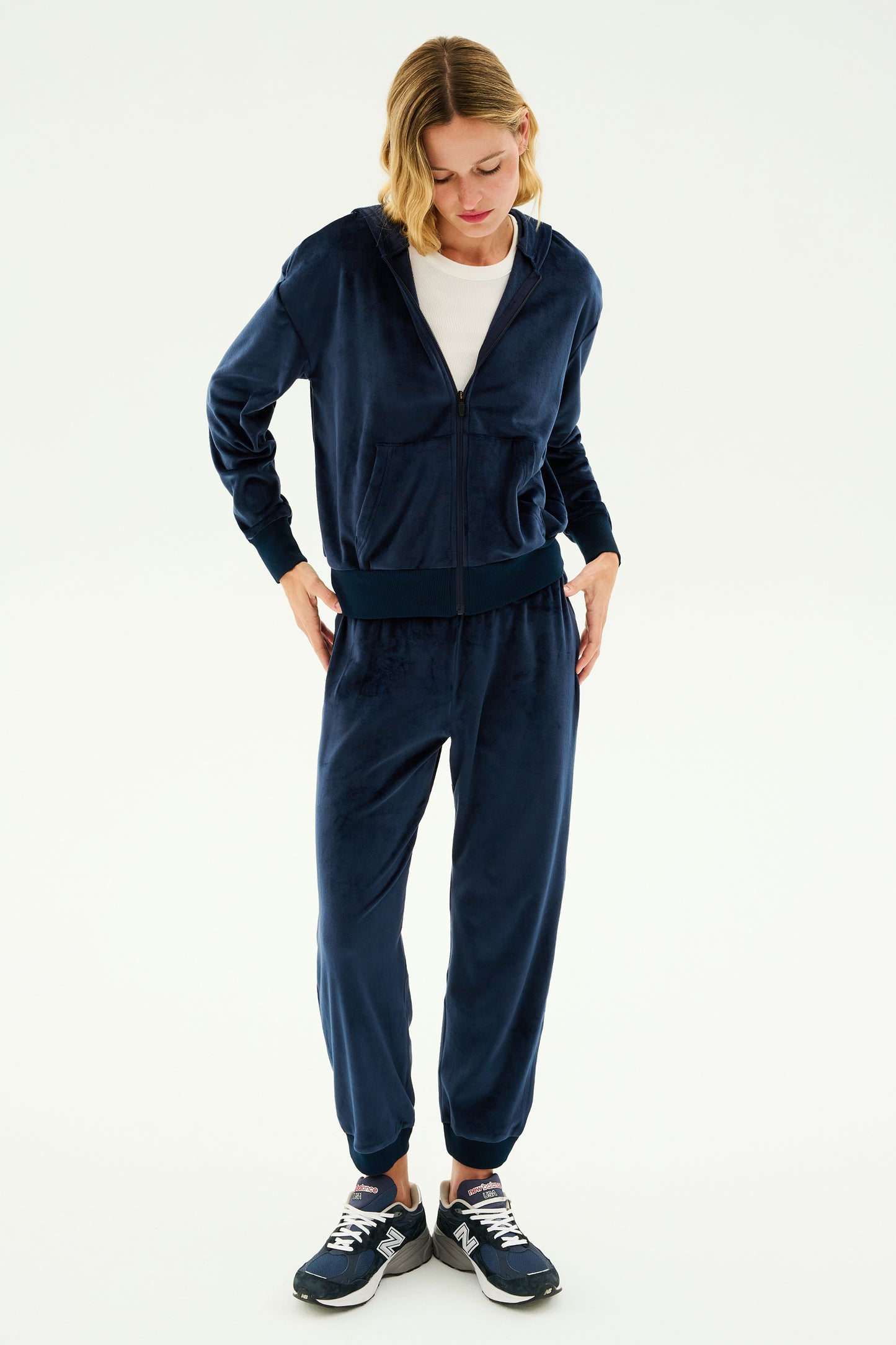 A person is lounging in an indigo Rhys Velour Jogger tracksuit from SPLITS59, paired with a white t-shirt and black sneakers, standing against a plain white background.