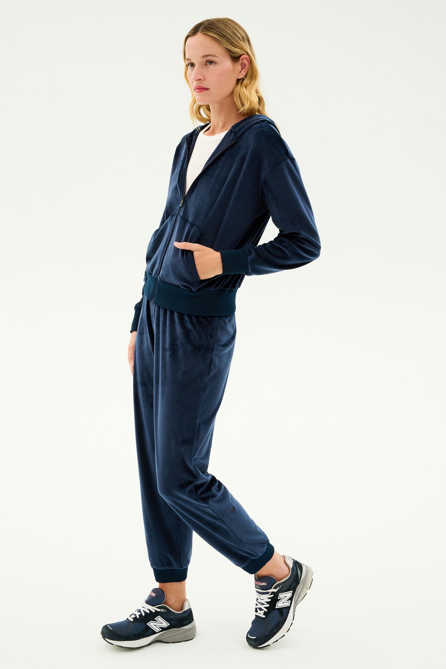 A woman wearing the SPLITS59 Rhys Velour Jogger - Indigo and a white top with matching navy blue sneakers, perfect for casual wear or lounging, stands against a plain background.