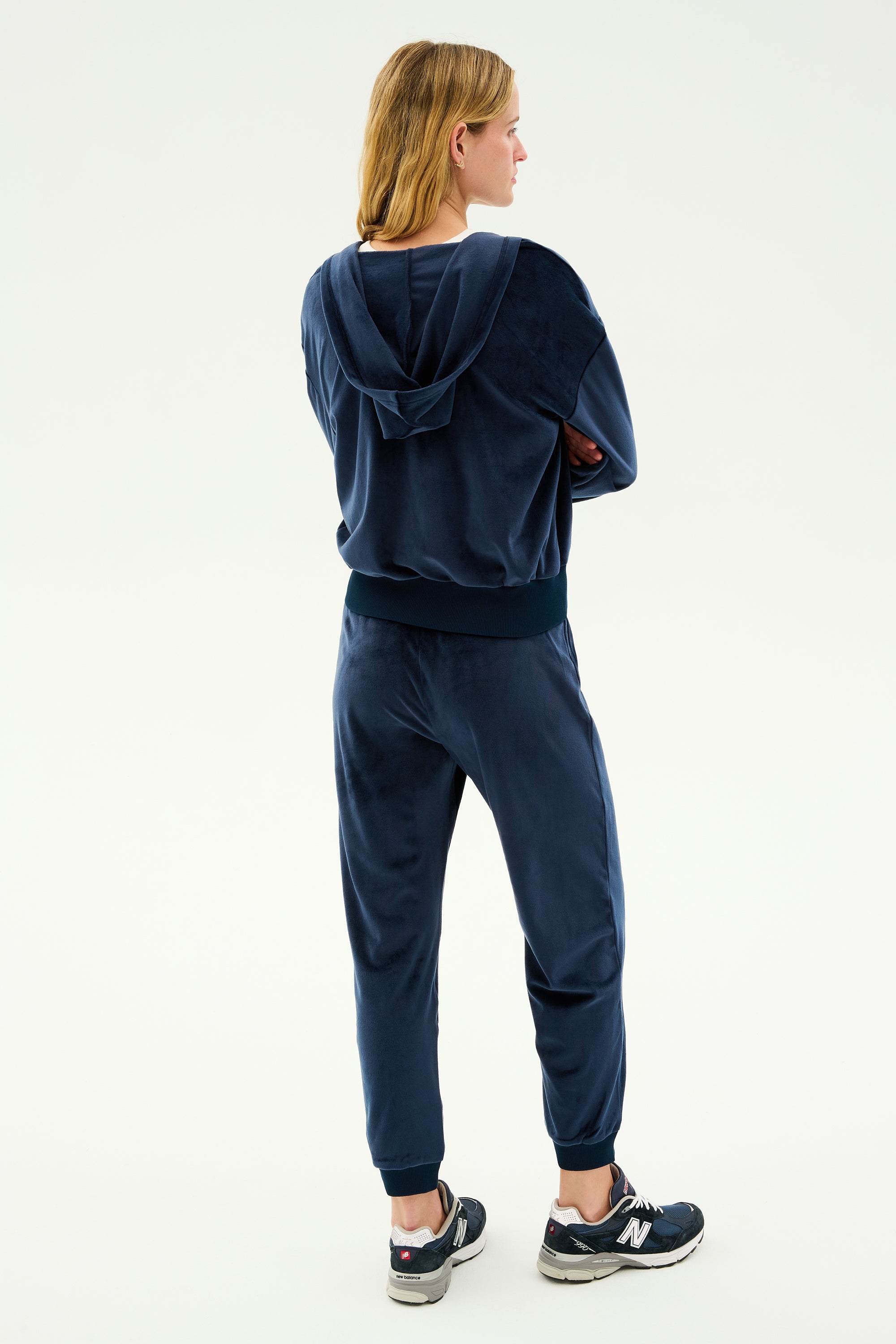 The individual with long blond hair is standing with arms crossed, back to the camera, wearing a navy blue hoodie and matching SPLITS59 Rhys Velour Jogger in Indigo, paired with black New Balance sneakers.