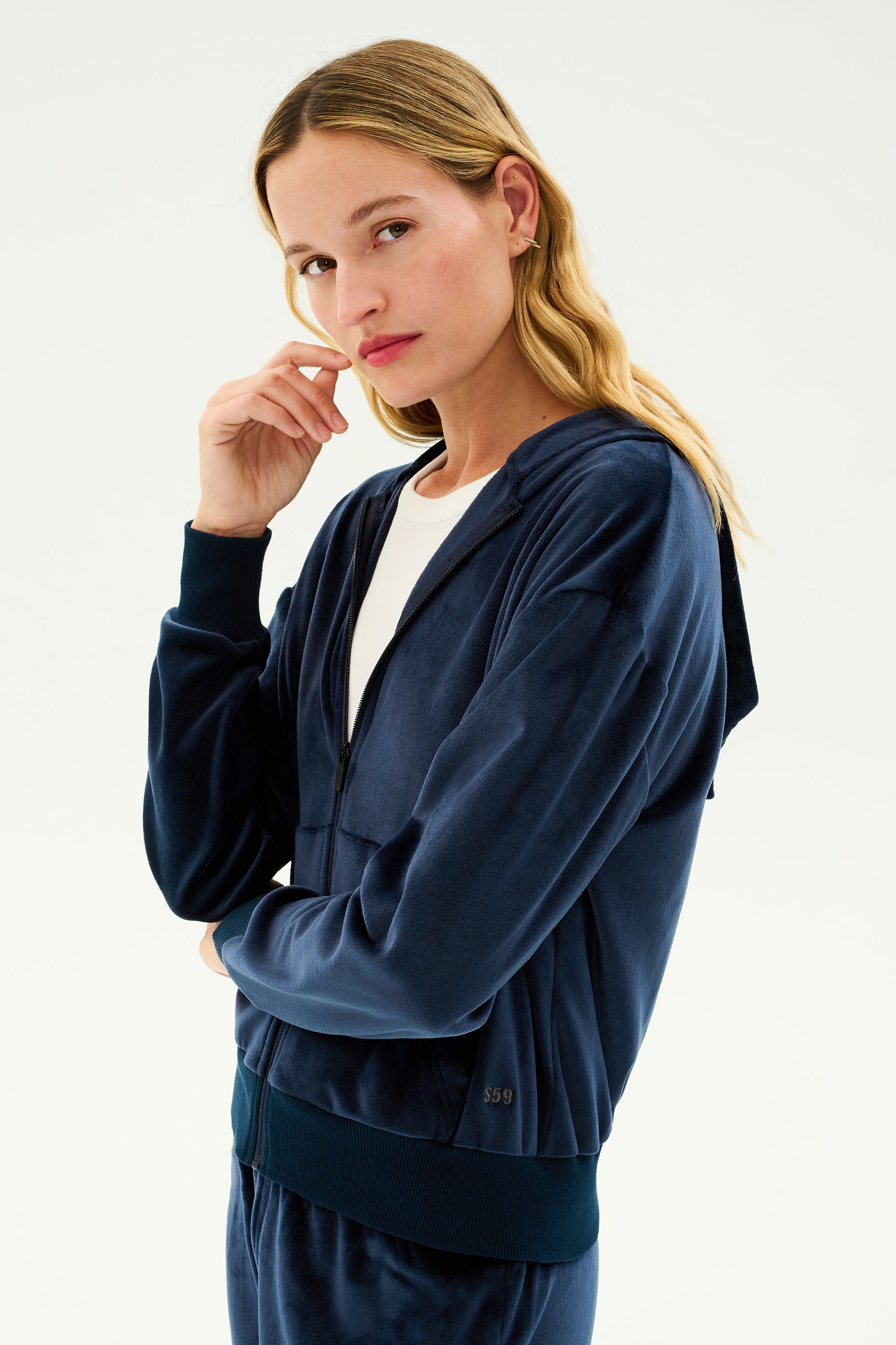 A person with long blond hair wearing a comfortable Rhys Velour Zip Up Hoodie in indigo and a white shirt stands with one hand near their face, looking at the camera.