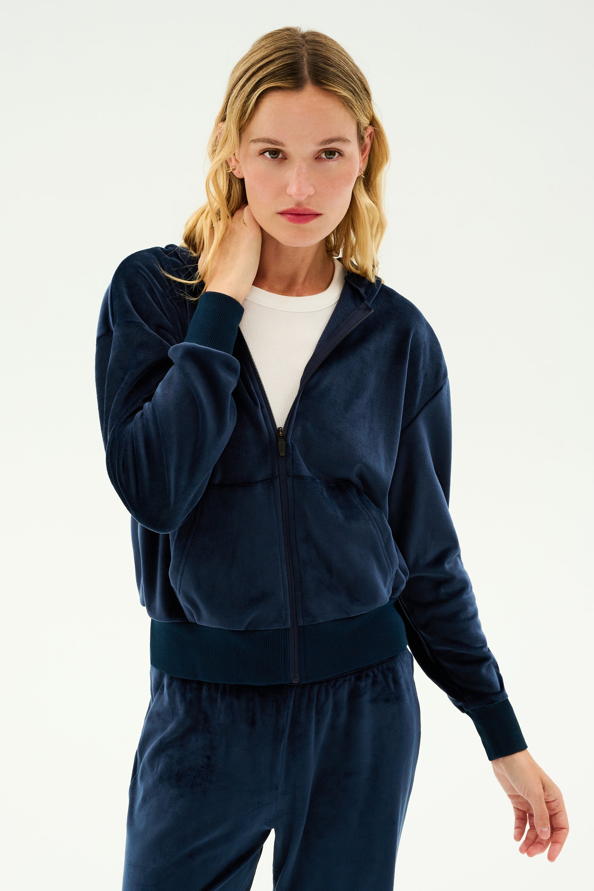 A person with long blonde hair is wearing the SPLITS59 Rhys Velour Zip Up Hoodie in Indigo and a white top, perfect for warming up and cooling down, standing against a plain background.