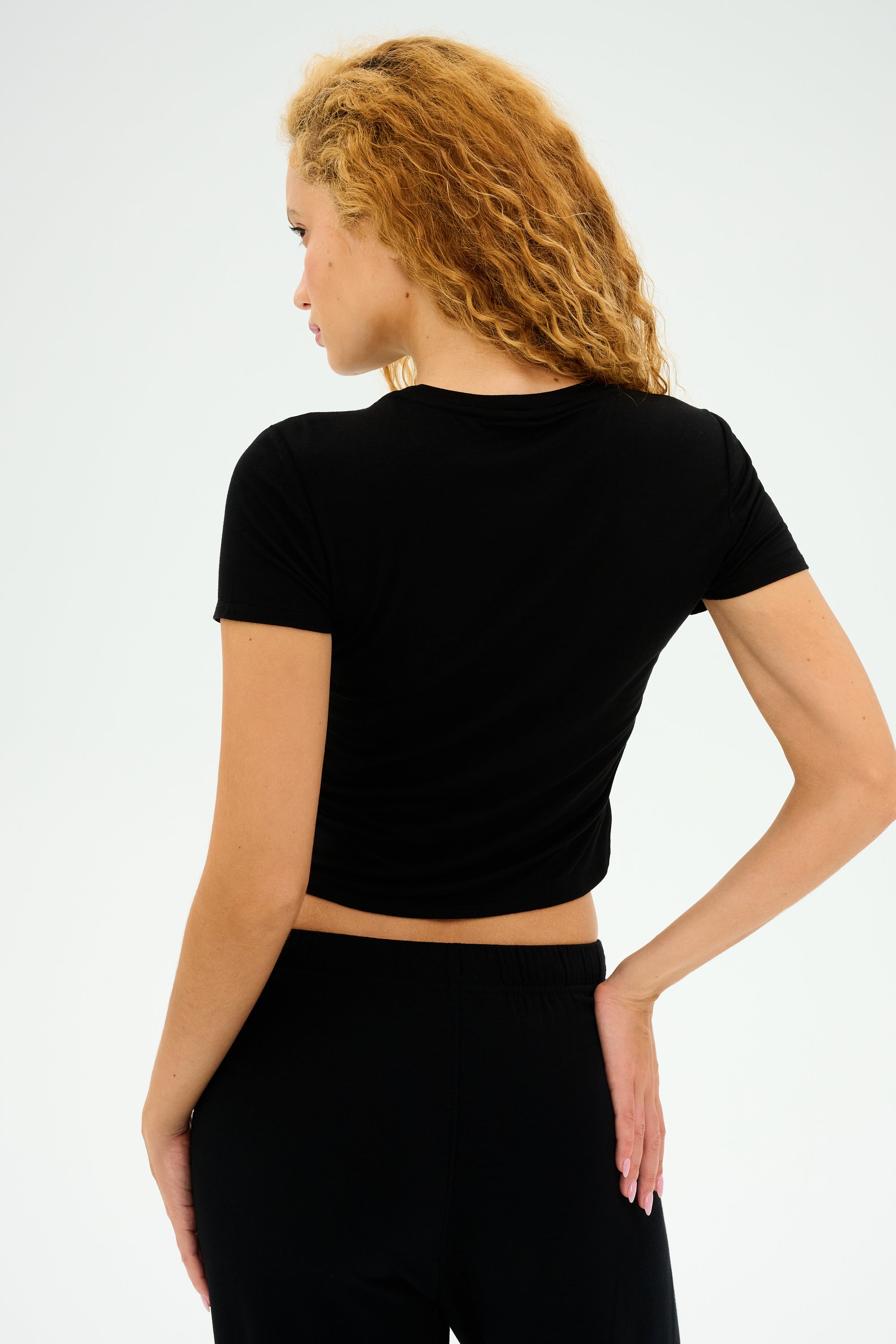 A curly-haired individual, wearing the SPLITS59 Jennie Jersey Tee in black and matching pants, stands against a white background with one hand on their hip.