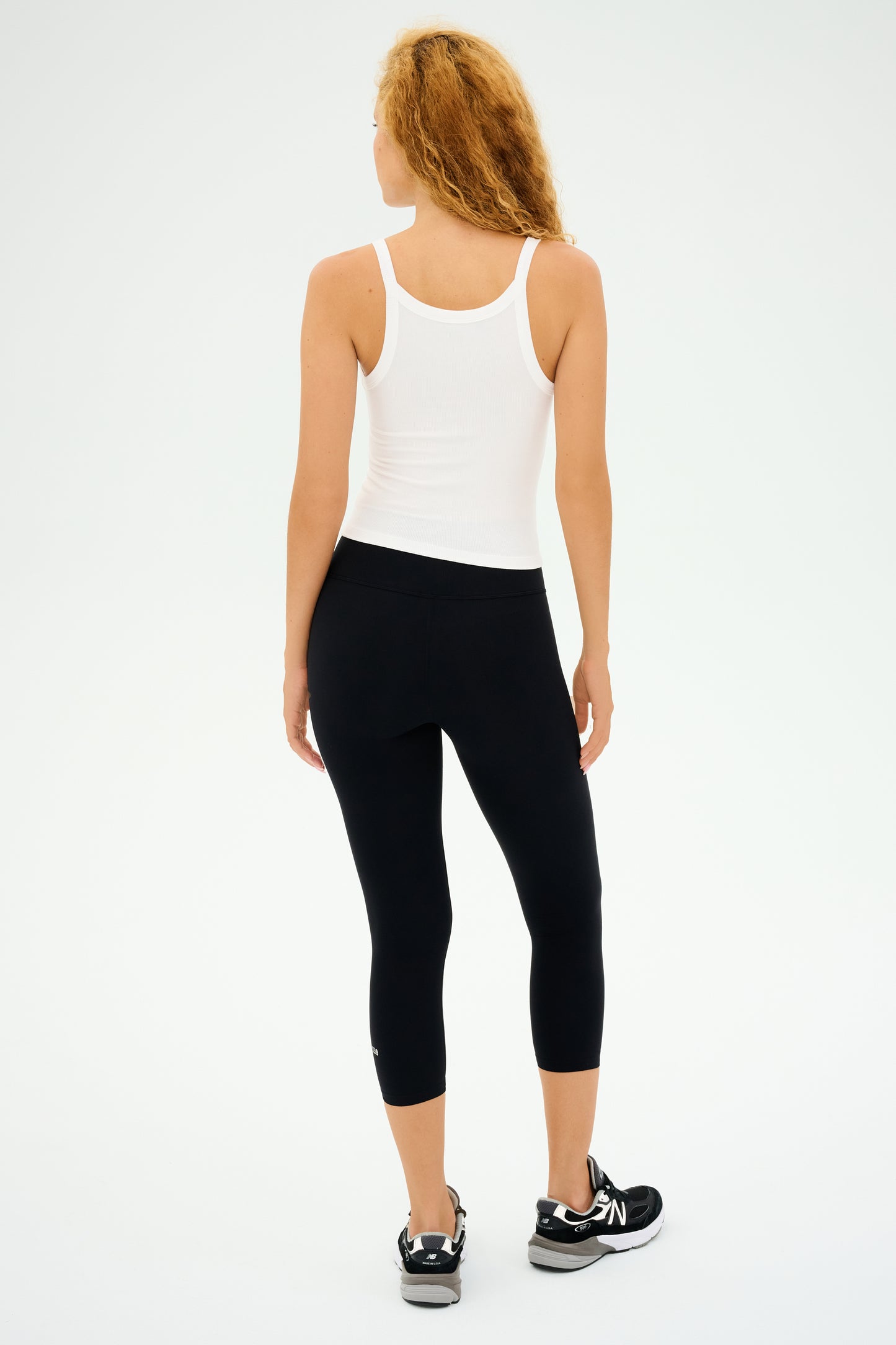 A person with long, curly hair wears the SPLITS59 Charlie Rib Tank in white, black leggings, and sneakers while standing and looking away against a plain background.