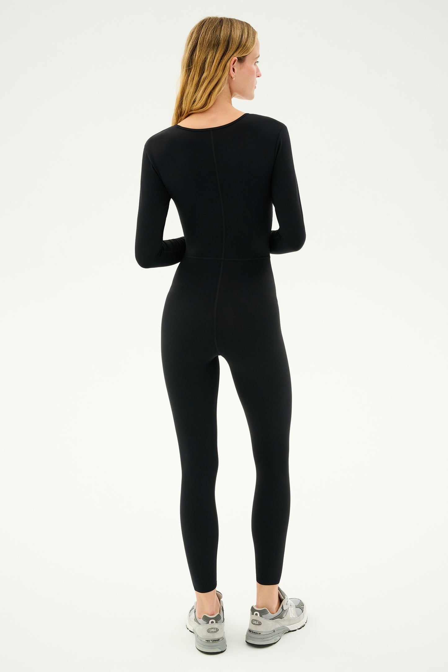 A person wearing the Chloe Airweight Jumpsuit in black from SPLITS59 and silver sneakers stands facing away from the camera against a plain background.