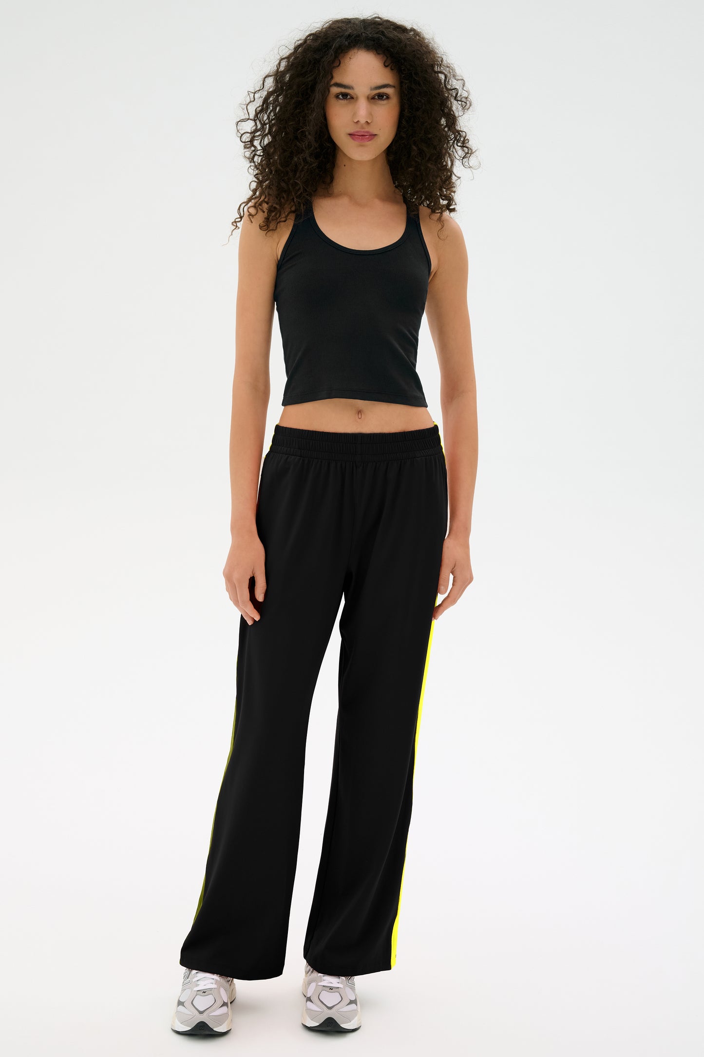 A person with curly hair stands wearing a black tank top, Maxie Rigor Track Pant in Black/Chartreuse by SPLITS59, and white sneakers against a white background.