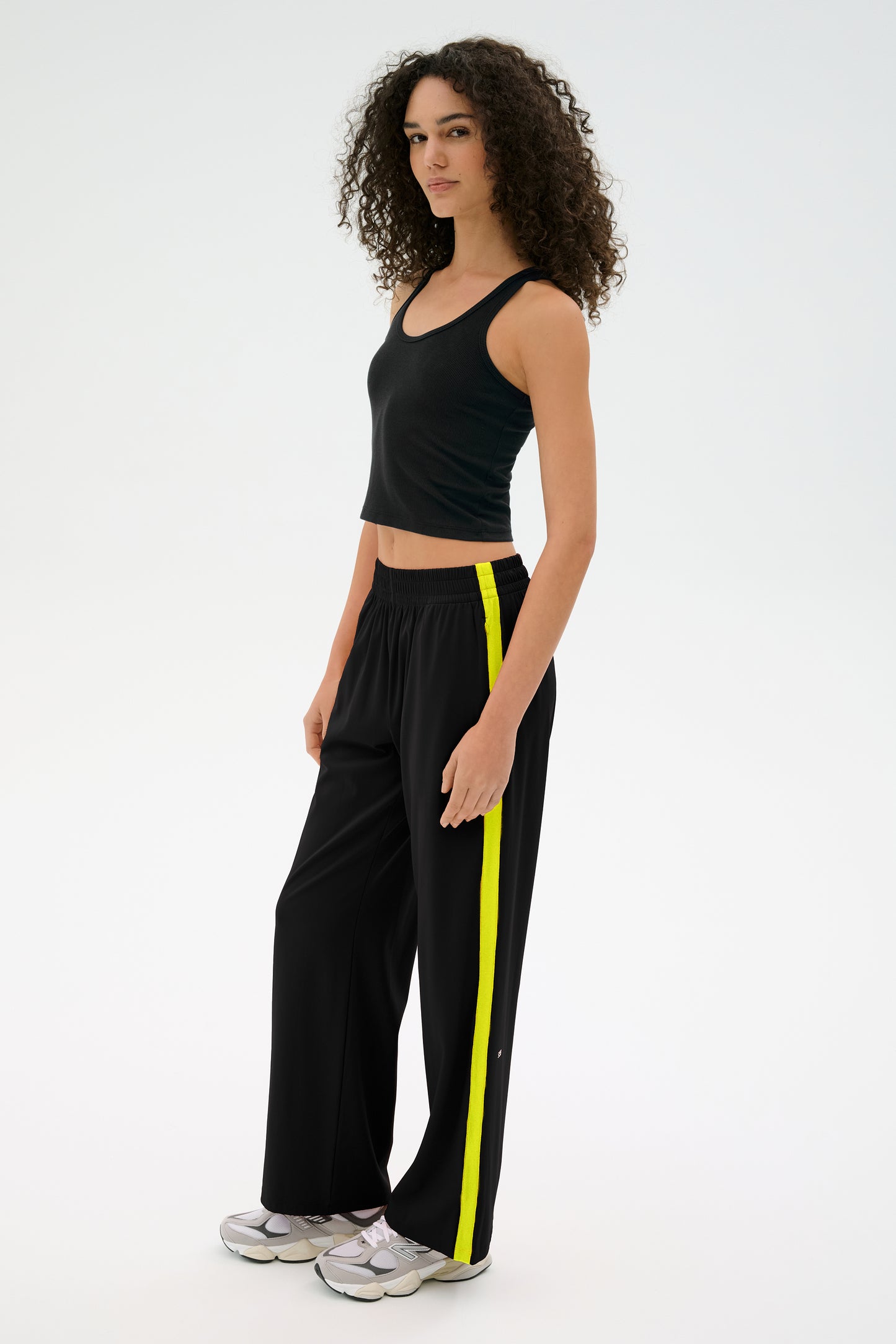 A person with curly hair stands against a plain background, sporting a black tank top and SPLITS59 Maxie Rigor Track Pant in Black/Chartreuse, complemented by sneakers.