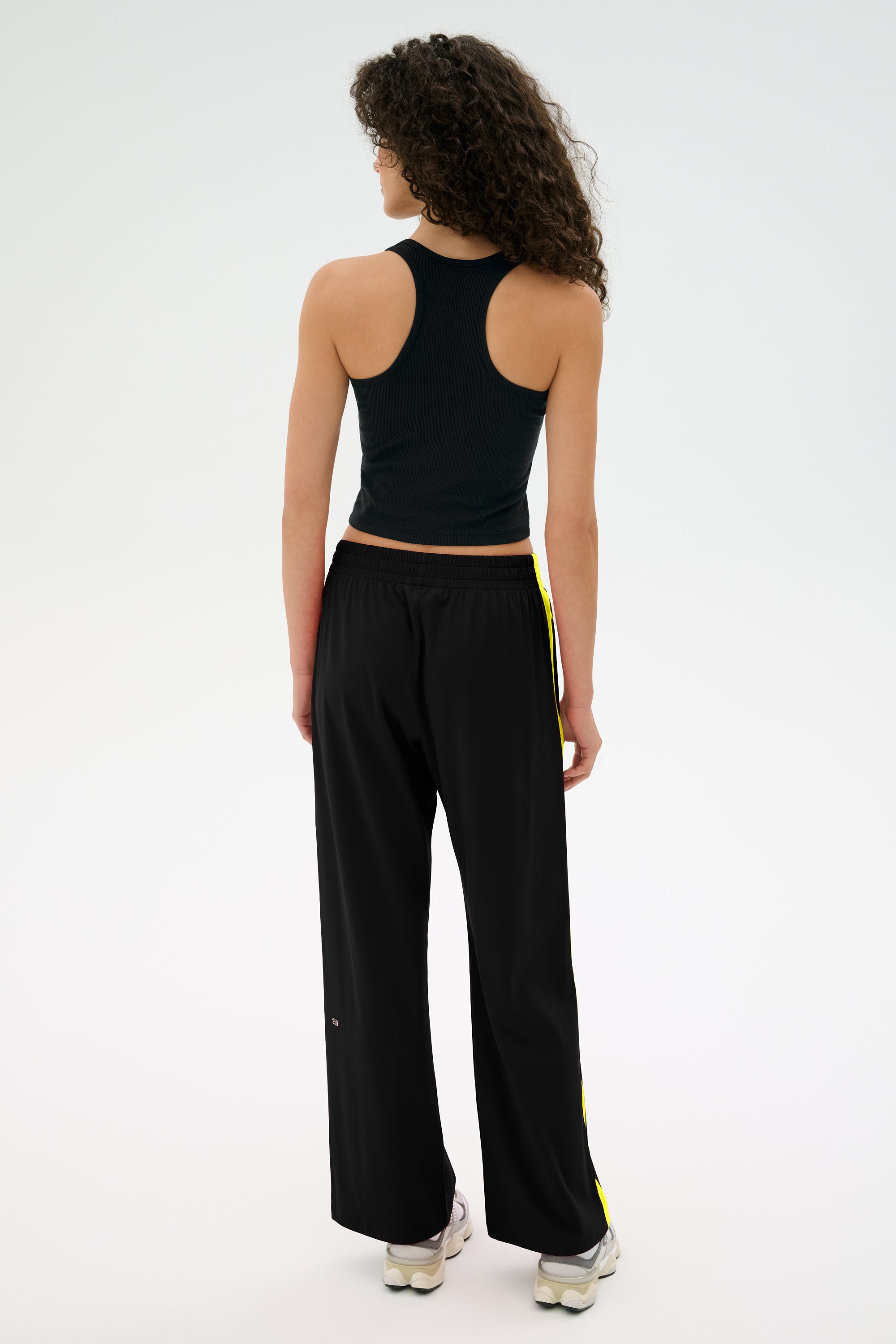 A person with curly hair is wearing a black tank top and SPLITS59's Maxie Rigor Track Pant in Black/Chartreuse, featuring yellow stripes, facing away.