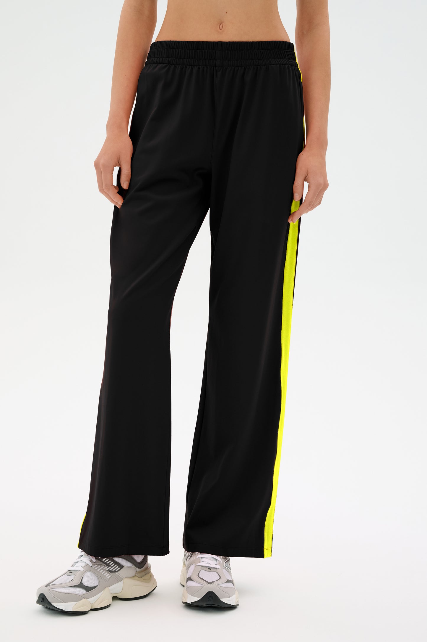 A person is dressed in SPLITS59's Maxie Rigor Flare Track Pant, which features black fabric with yellow side stripes, and pairs it with white sneakers against a neutral backdrop.