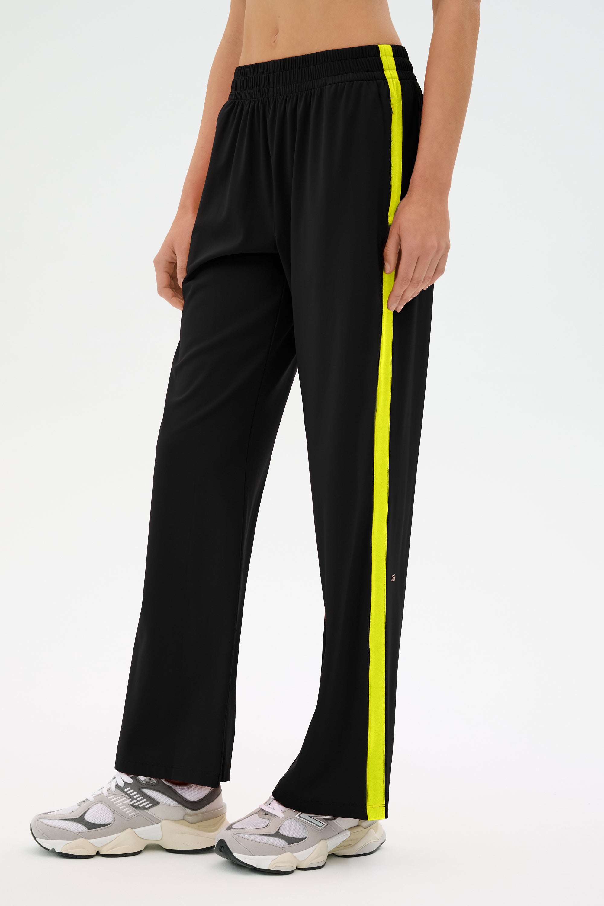 A person is wearing the SPLITS59 Maxie Rigor Track Pant in Black/Chartreuse, showcasing a wide leg silhouette with a bold yellow side stripe, effortlessly paired with white sneakers.