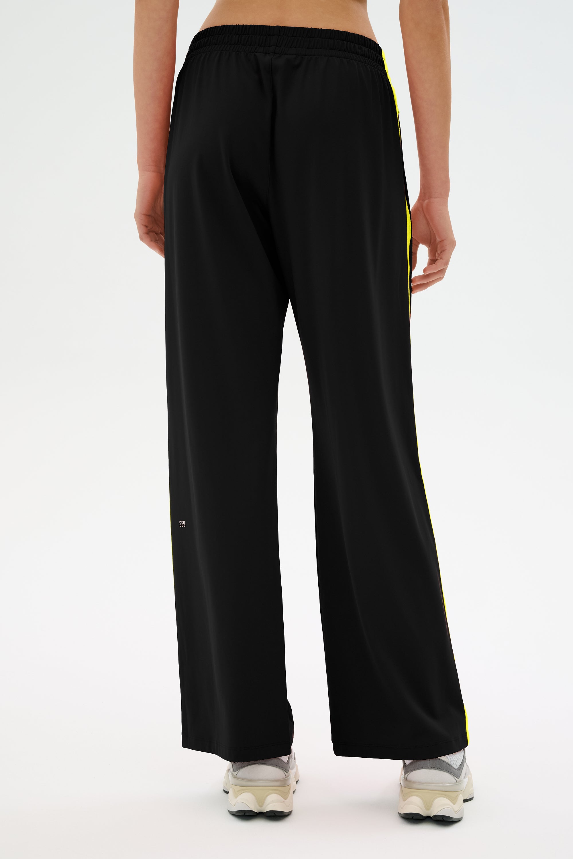 Individual wearing SPLITS59's Maxie Rigor Track Pant in Black/Chartreuse with white sneakers, captured from behind.