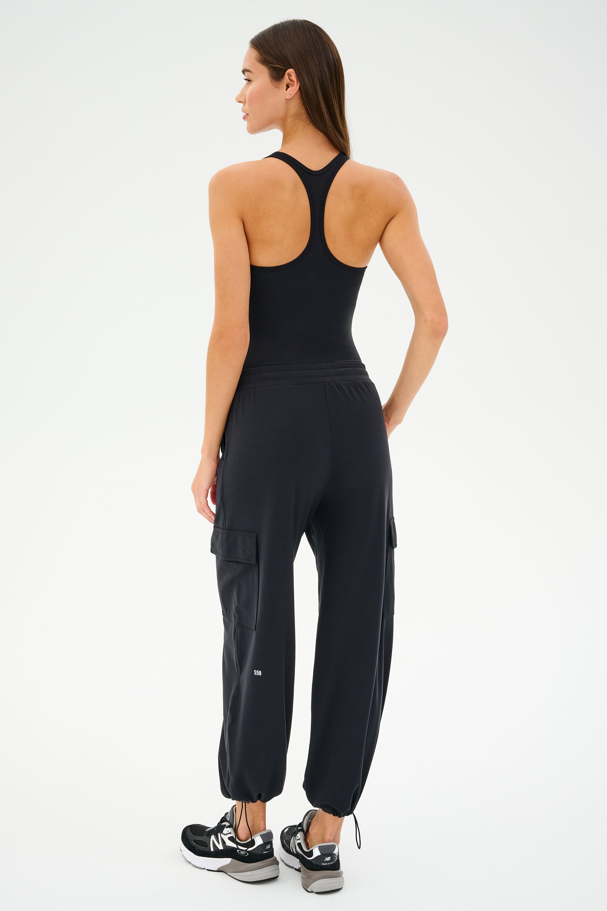 Dressed in SPLITS59's Nico Airweight Cargo Pant - Black and a black tank top, a person stands facing away from the camera in sleek black sneakers.