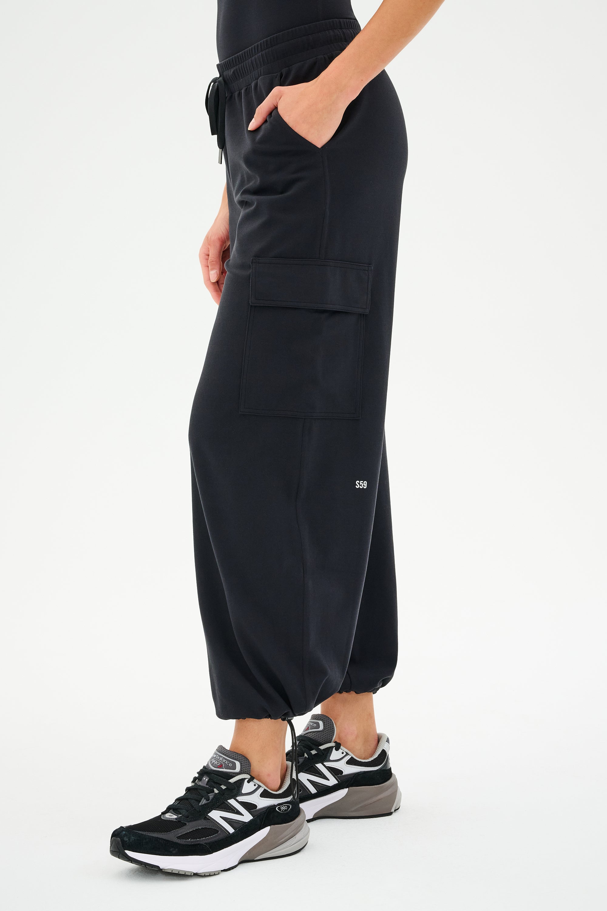 A person wearing SPLITS59's Nico Airweight Cargo Pant in black and New Balance sneakers stands on one foot with hands in pockets against a plain background.