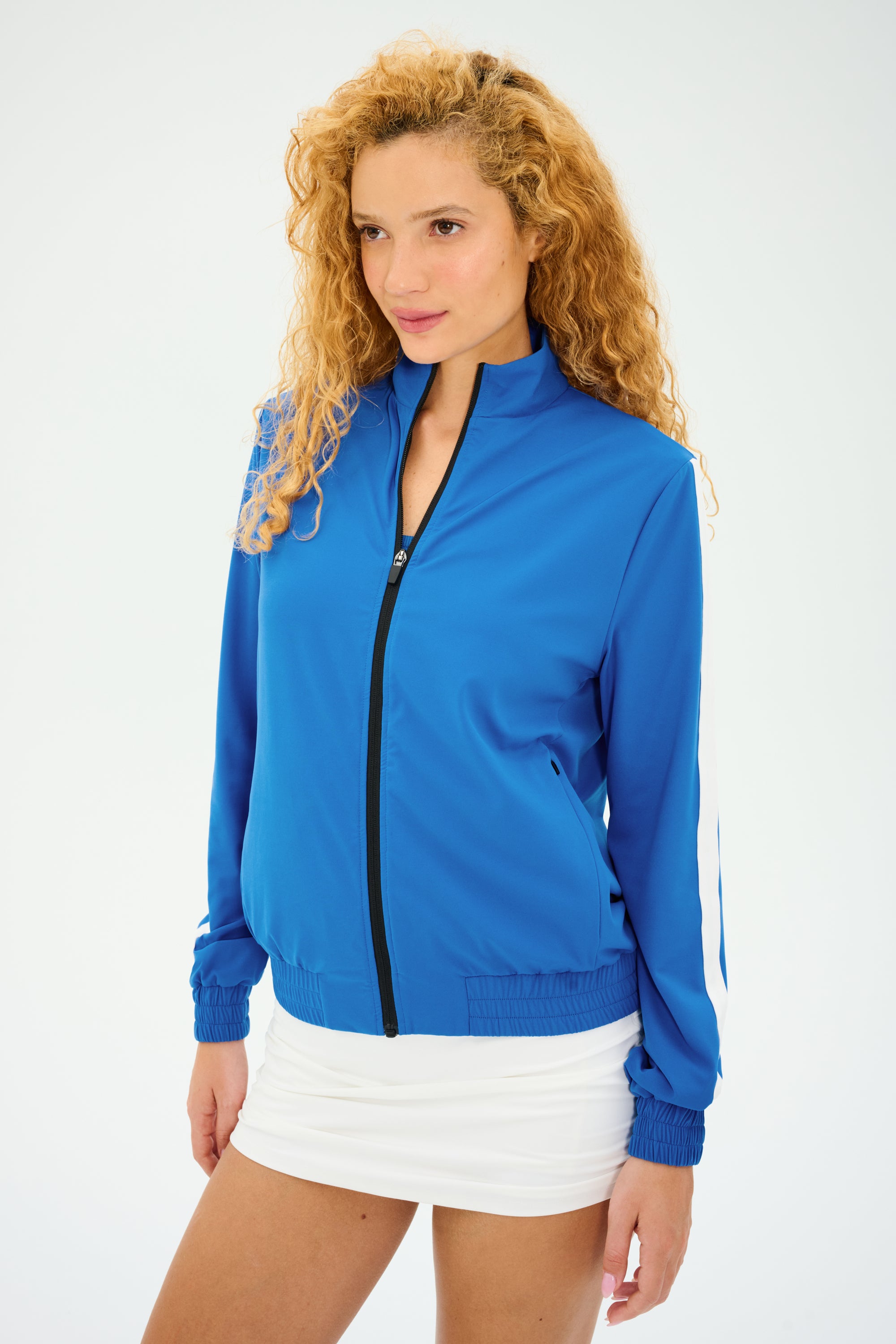 The individual is wearing a SPLITS59 Max Rigor Track Jacket in Classic Blue/White and a white skirt, looking thoughtfully to the side against a plain backdrop.