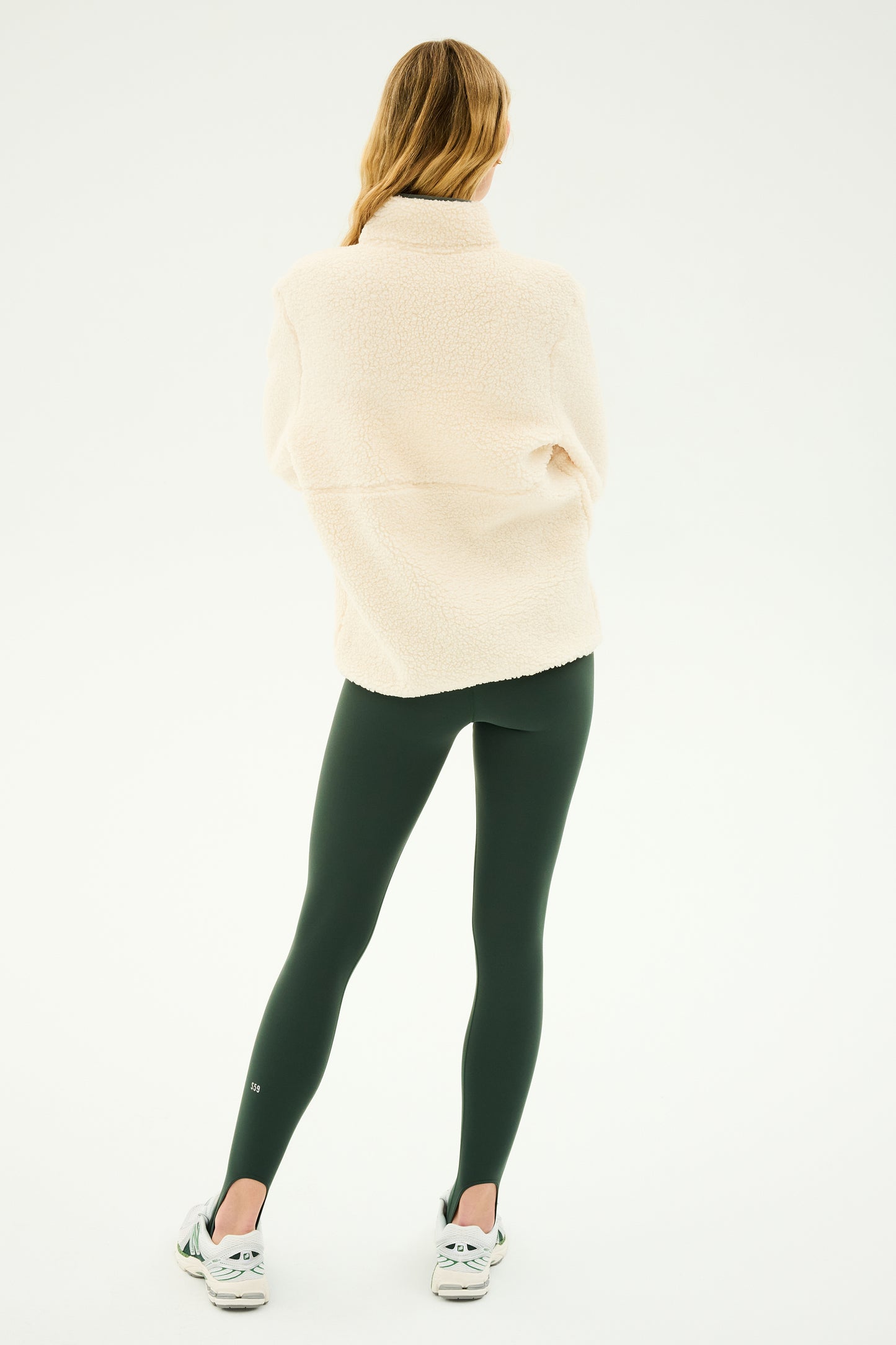 A person with long hair is standing with their back to the camera, wearing SPLITS59's Libby Sherpa Half Zip in Creme/Military along with dark green leggings and white sneakers—ready for winter workouts or perhaps some yoga practice.