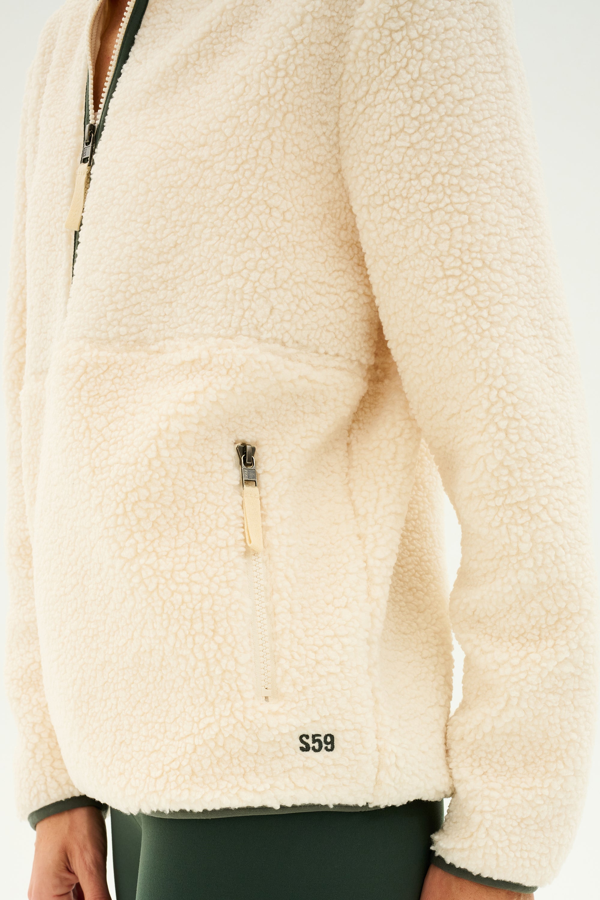 A person wearing the SPLITS59 Libby Sherpa Half Zip in Creme/Military, featuring a front pocket and "S59" embroidered near the hem. Only the upper torso and arms are visible in the image.