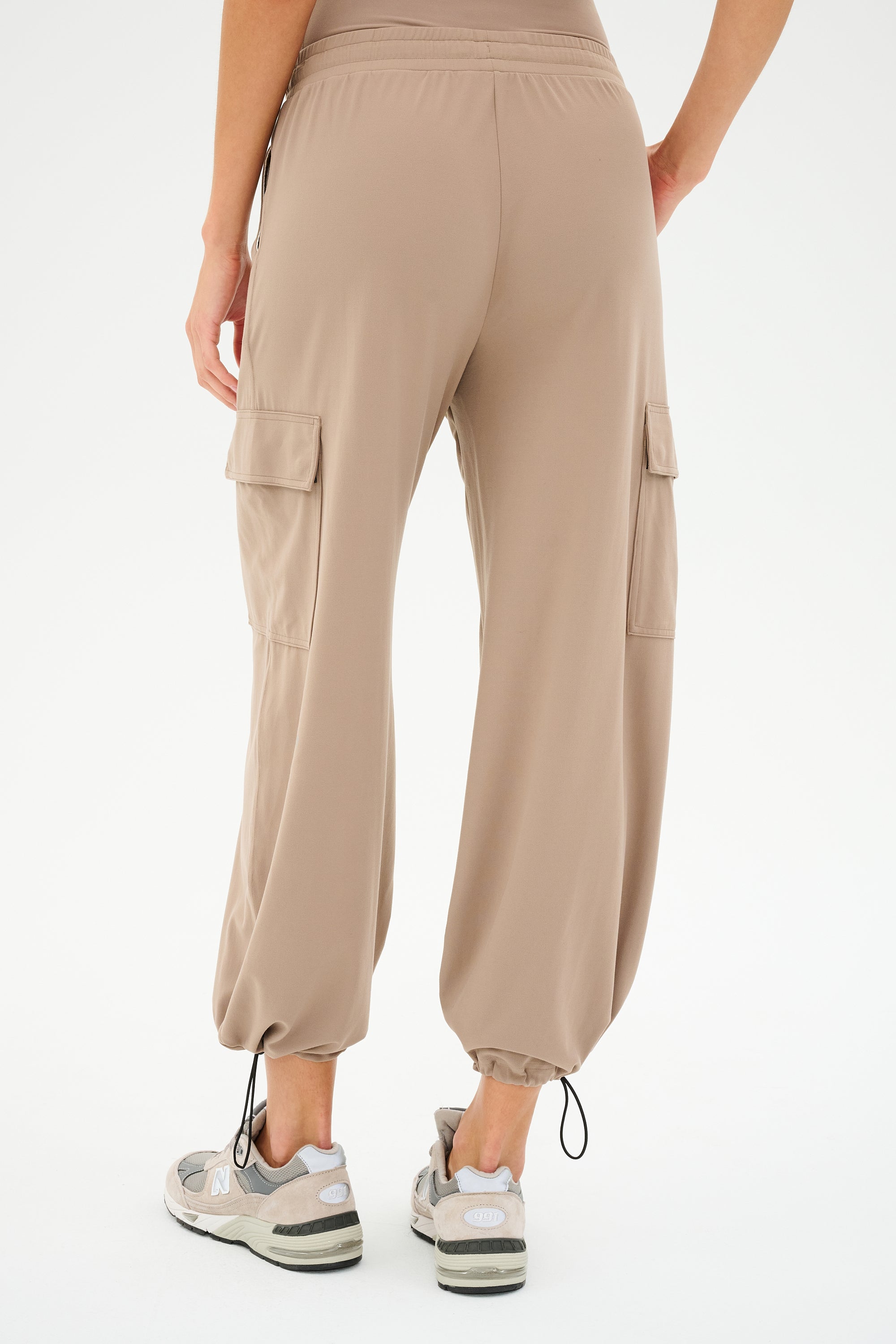 A person is seen from behind wearing SPLITS59's Nico Airweight Cargo Pant in Latte, featuring drawstring cuffs, and gray sneakers against a white backdrop.