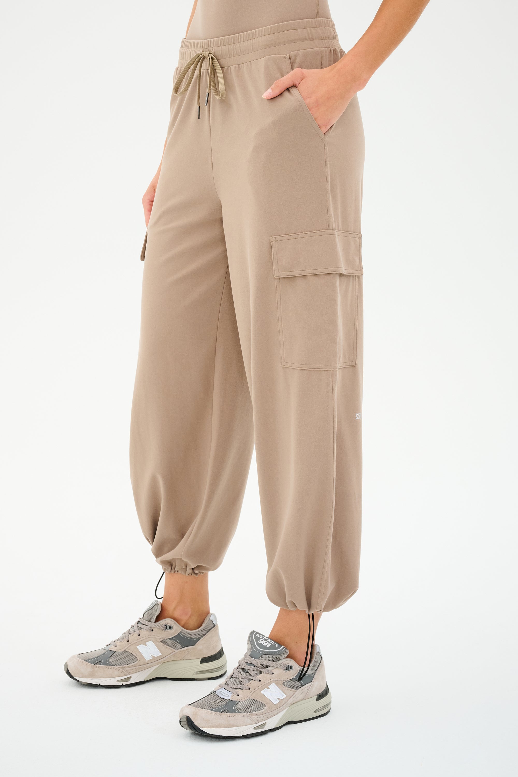 A person wearing SPLITS59's Nico Airweight Cargo Pant in Latte paired with gray sneakers, standing with a hand in one pocket.