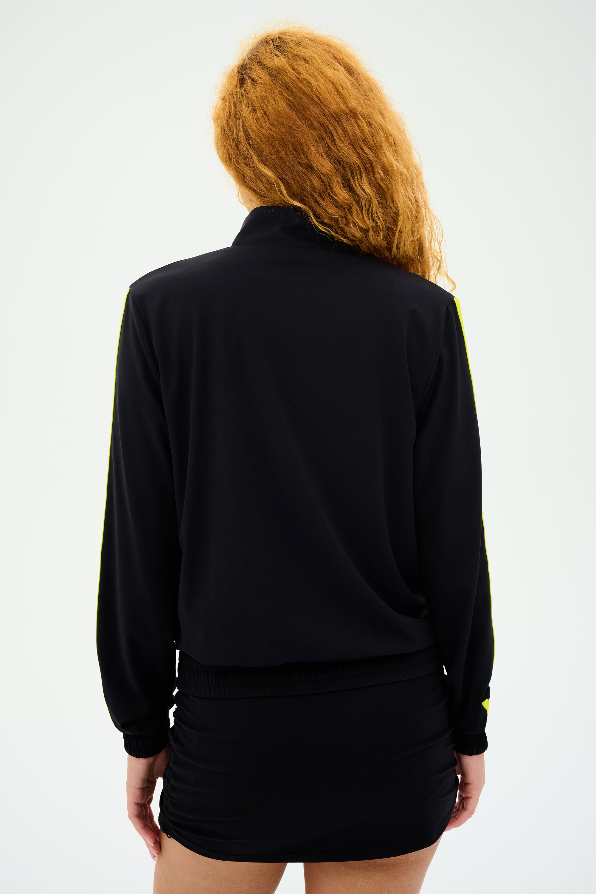 Individual with long red hair wearing the SPLITS59 Max Rigor Track Jacket in Black/Chartreuse and skirt, facing away.