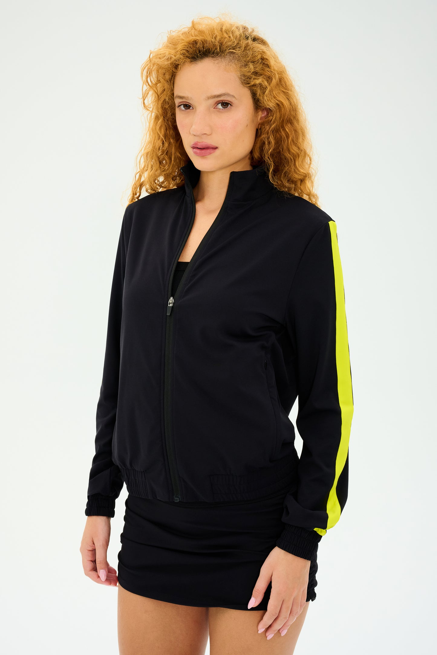 A curly-haired individual wears the SPLITS59 Max Rigor Track Jacket in Black/Chartreuse with a chic black dress, set against a simple backdrop.