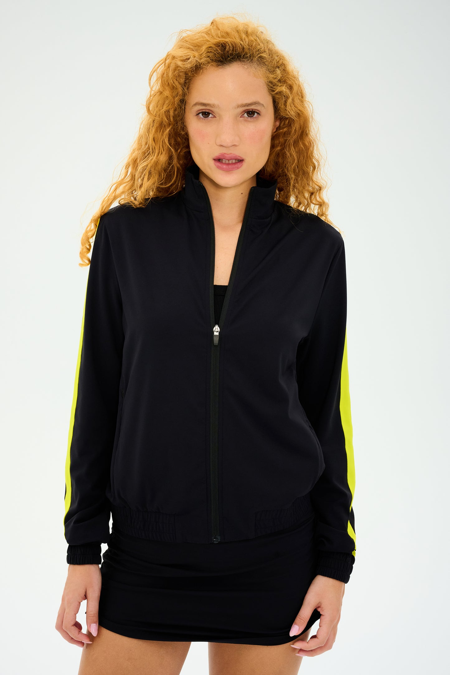 A person with long curly hair confidently poses against a plain background, wearing the stylish SPLITS59 Max Rigor Track Jacket in Black/Chartreuse and paired with a chic skirt.