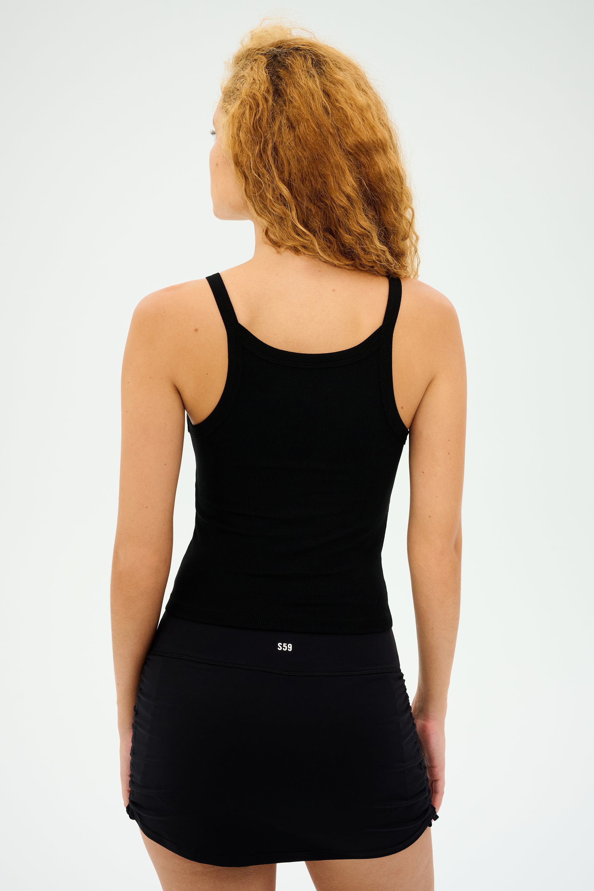 A curly-haired individual models the SPLITS59 Charlie Rib Tank in black, paired with a skirt and set against a neutral backdrop.