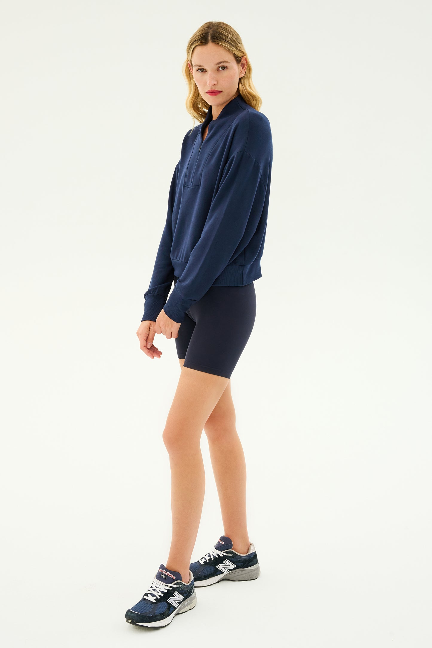 A person stands wearing the Birdie Fleece Half Zip in Indigo from SPLITS59, black shorts, and navy sneakers against a plain background.