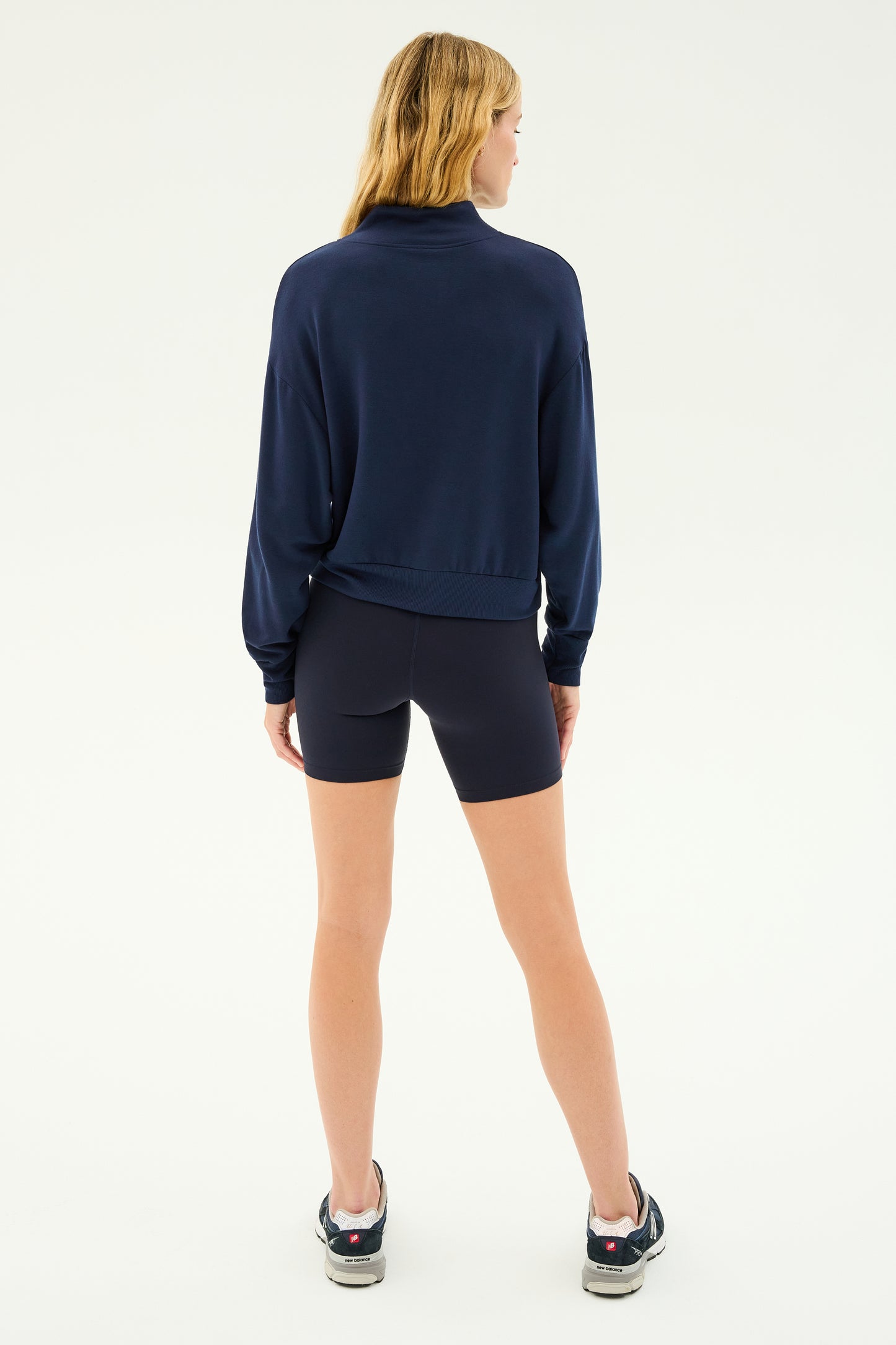 A person with long hair stands facing away, wearing the SPLITS59 Birdie Fleece Half Zip in Indigo, paired with black athletic shorts and sneakers, against a plain background.