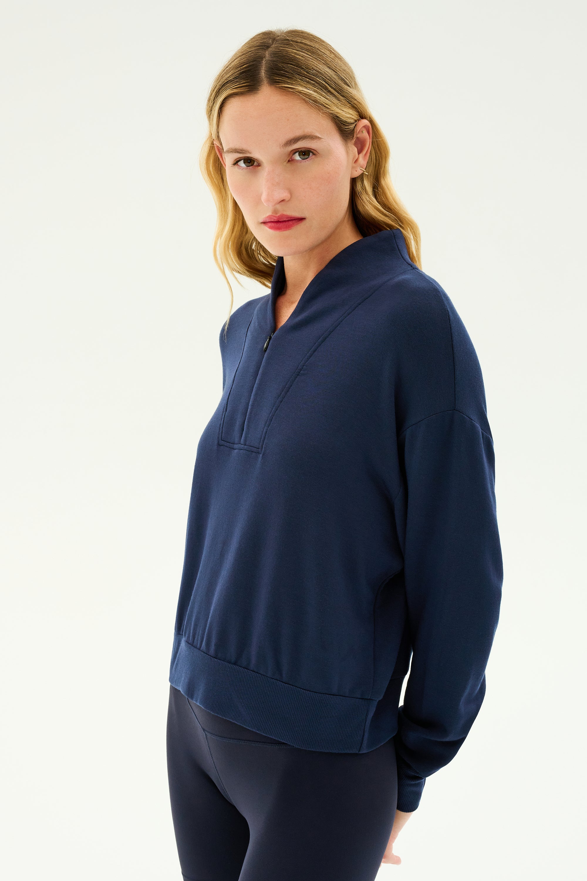 A person with long hair wearing the SPLITS59 Birdie Fleece Half Zip in Indigo stands against a plain white background, looking directly at the camera. The sleek design of the half-zip makes it perfect for winter workouts.