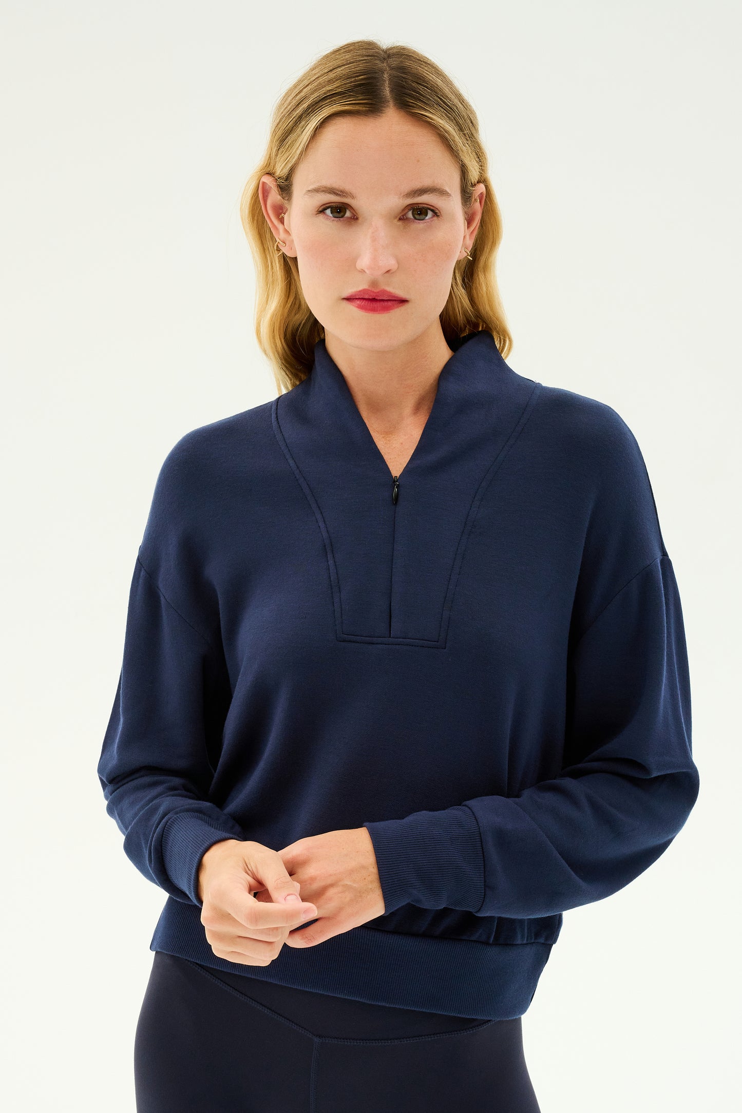 A person with light hair stands against a plain background, wearing the SPLITS59 Birdie Fleece Half Zip in indigo, featuring a cozy half-zip collar, perfect for winter workouts.