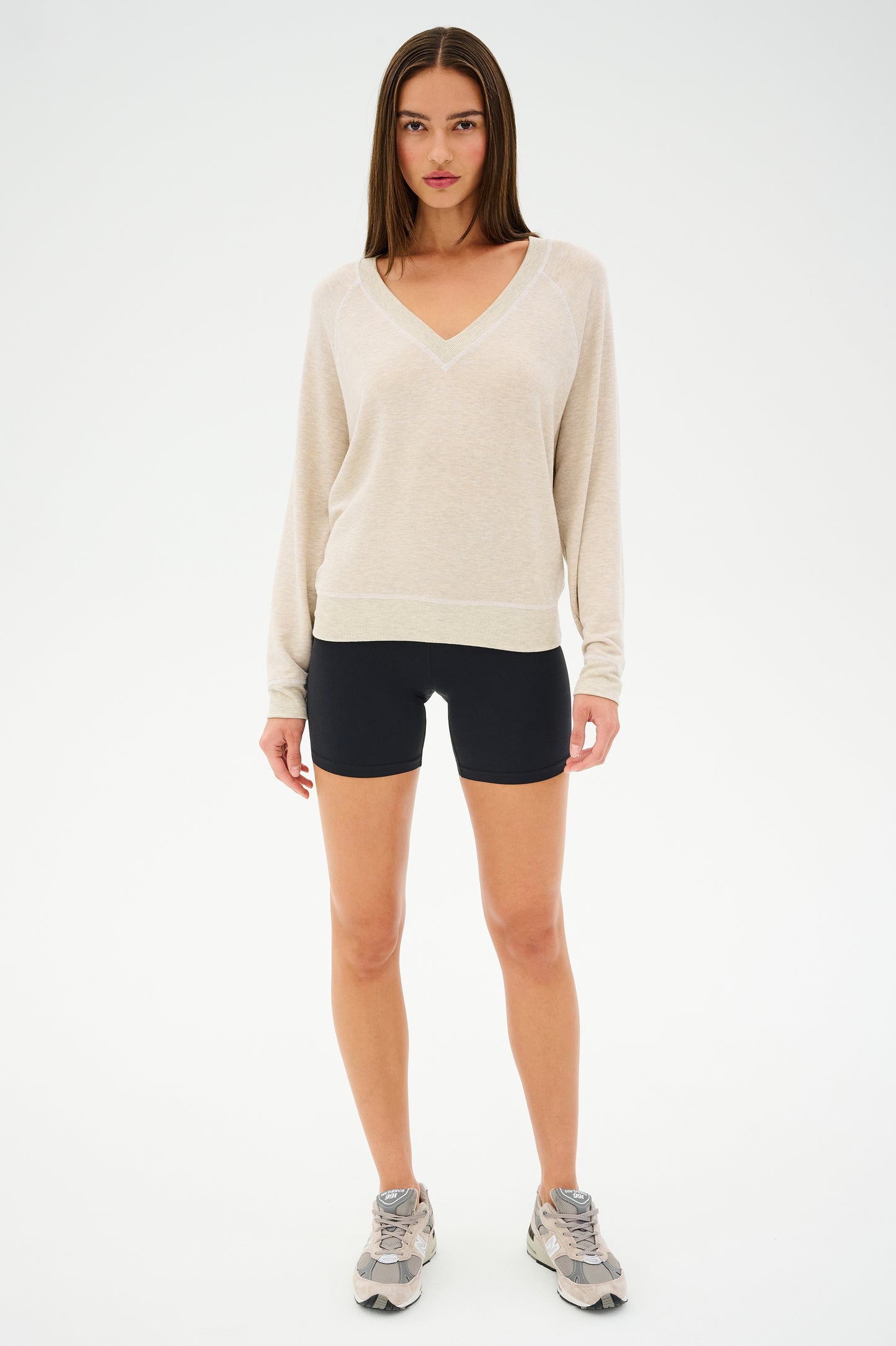 A person dressed in a SPLITS59 Bennie Fleece V-Neck Sweatshirt in Heather Oatmeal/White, black shorts, and gray sneakers stands against a plain white background.