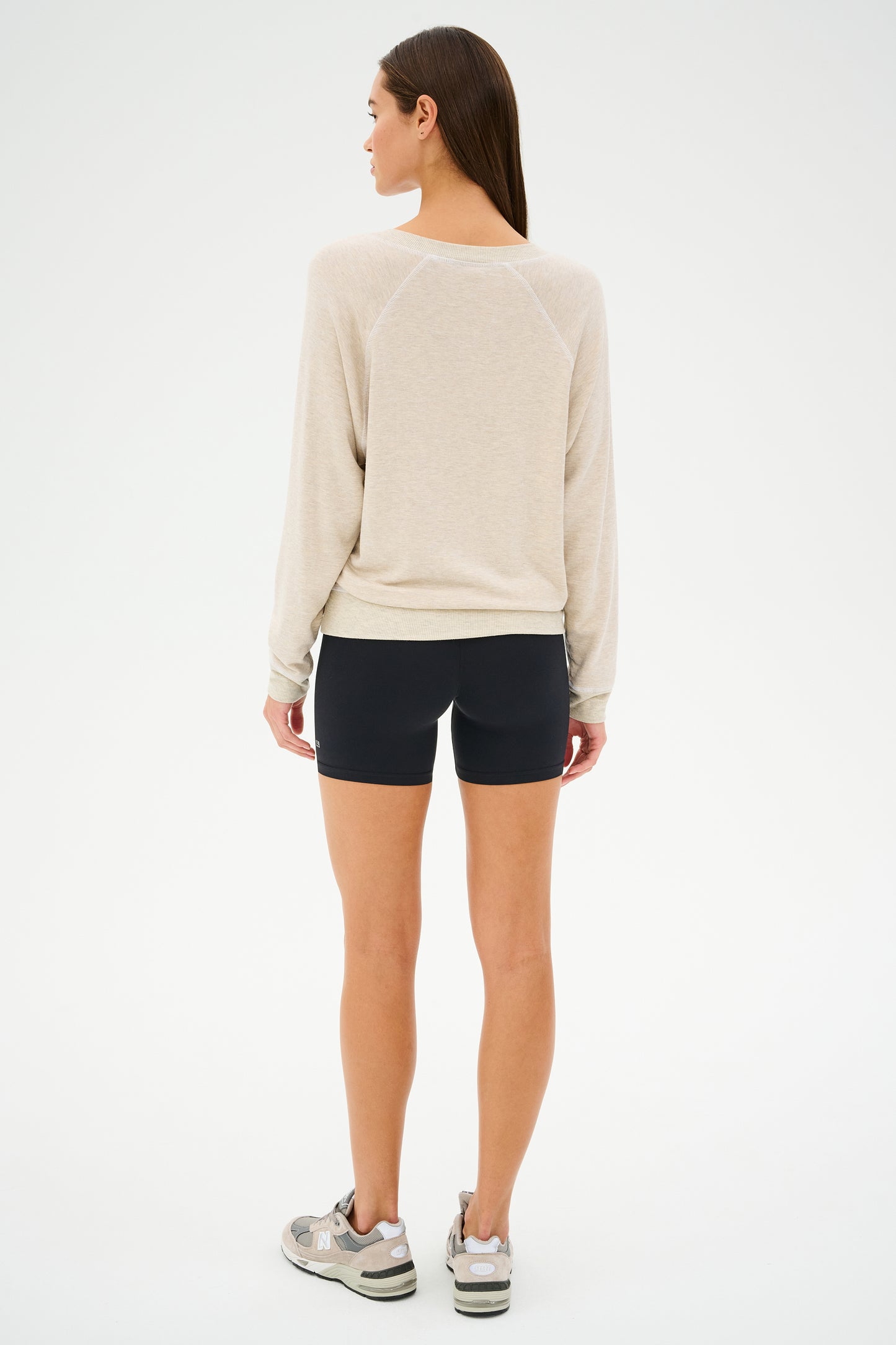 A person stands facing away, wearing SPLITS59 workout apparel, featuring a Bennie Fleece V-Neck Sweatshirt in Heather Oatmeal/White, black shorts, and sneakers against a plain background.