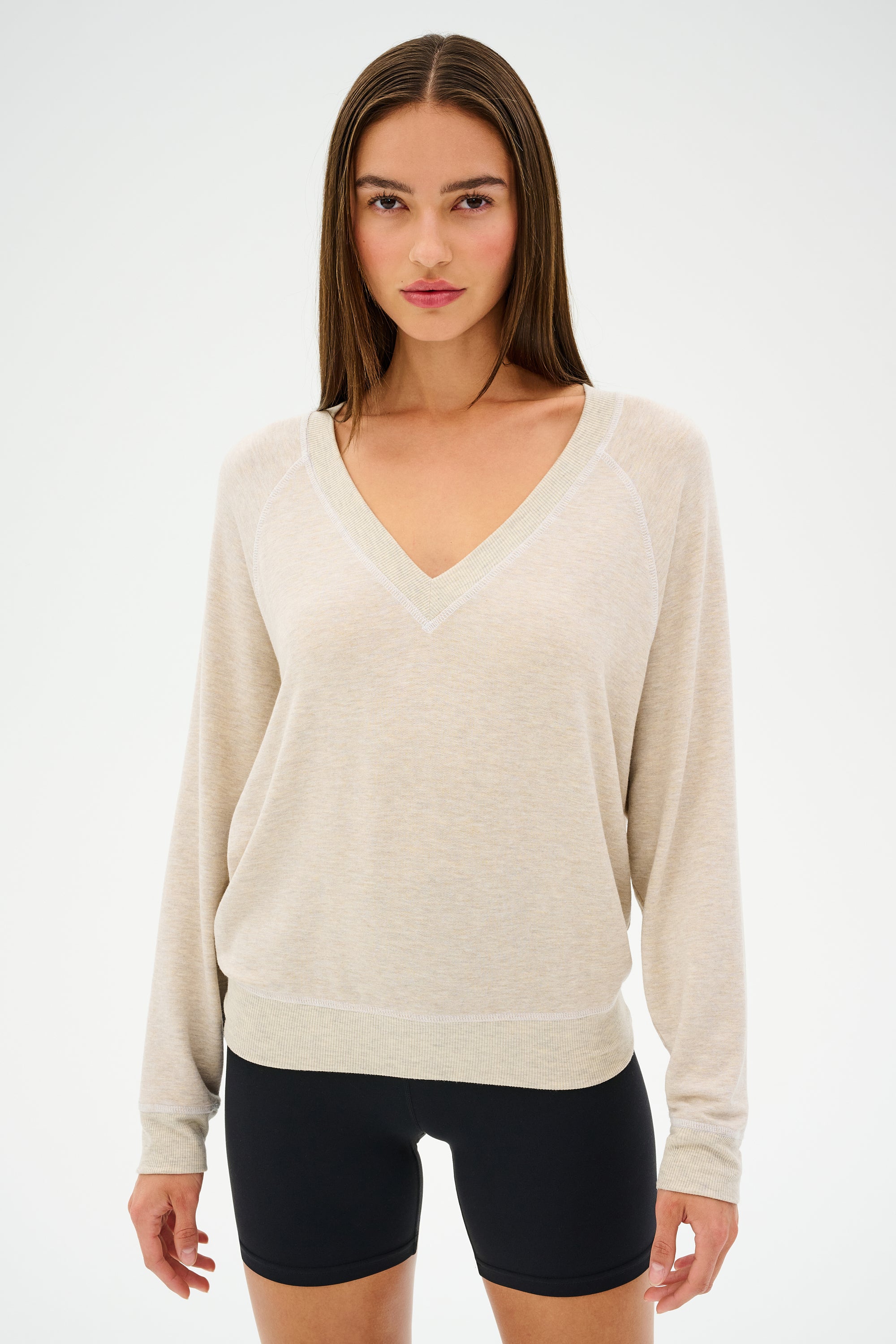 A woman in a SPLITS59 Bennie Fleece V-Neck Sweatshirt (Heather Oatmeal/White) and black shorts stands against a white background, showcasing stylish workout apparel.