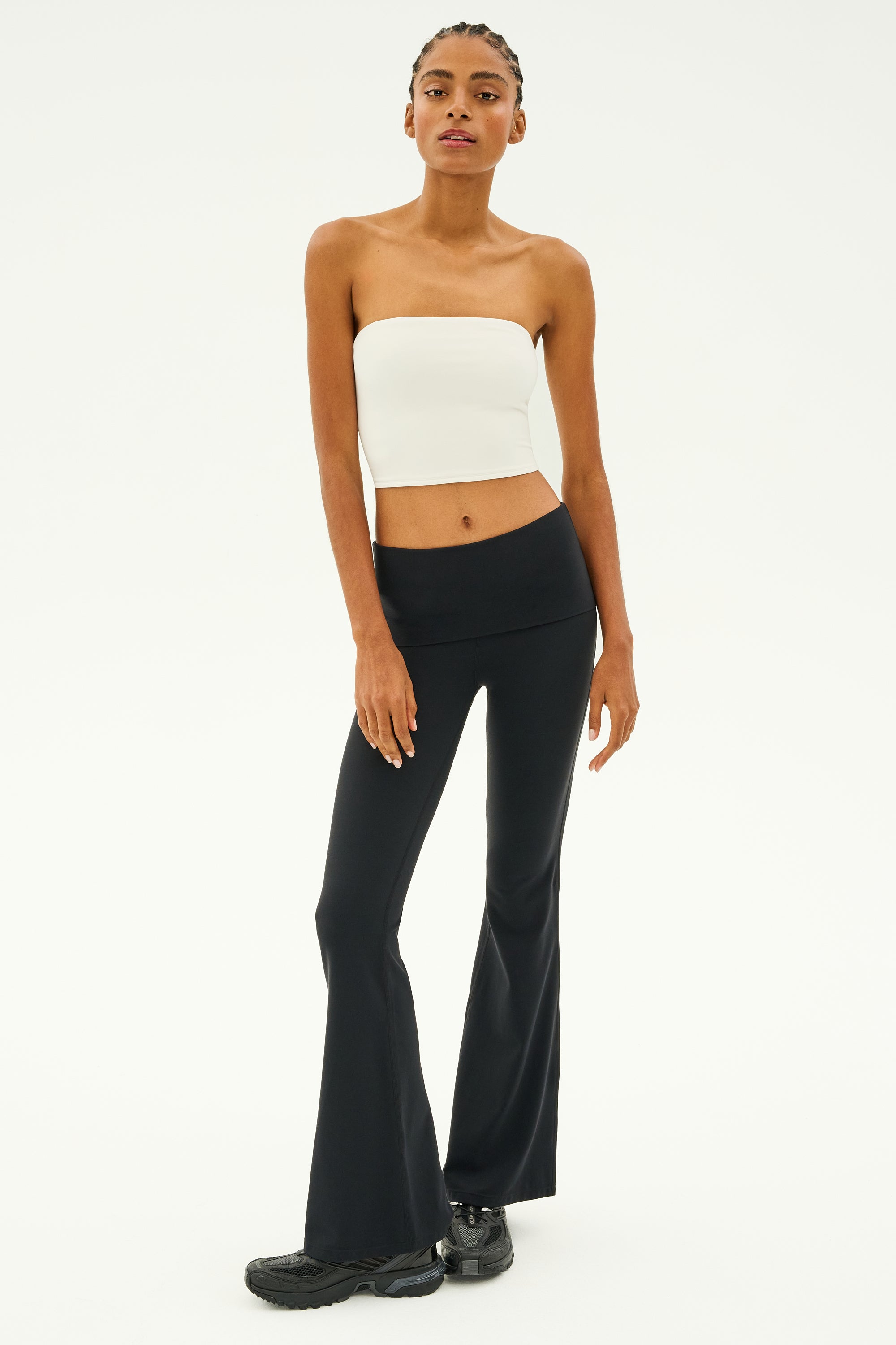 A person wearing a white strapless top and the Raquel Airweight Foldover Flare pants in black from SPLITS59, paired with black sneakers, stands against a plain background.