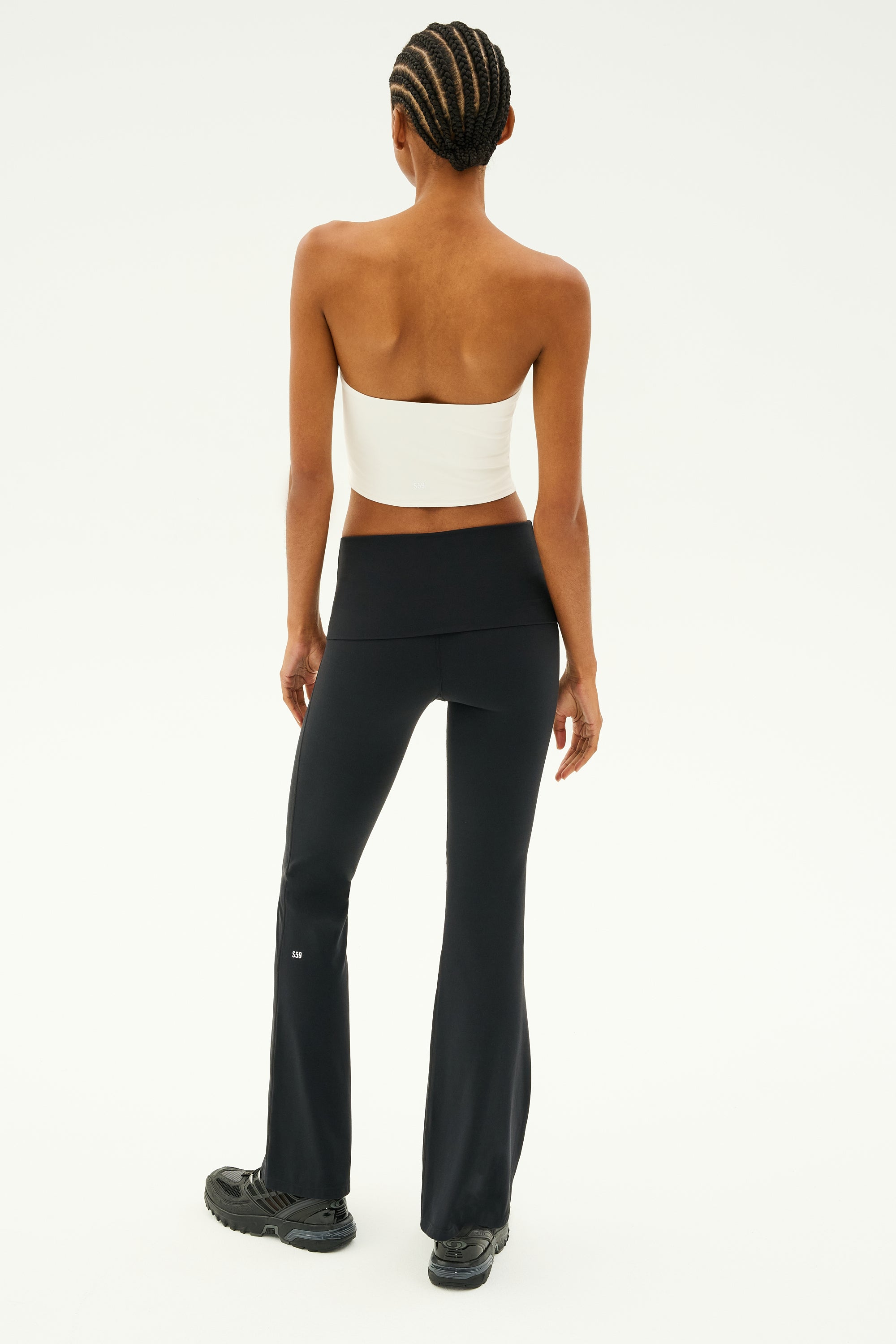 A person with braided hair stands facing away, wearing a white Hazel Airweight Crop Bandeau by SPLITS59 and black flared pants made from stretch fabric, paired with black shoes.