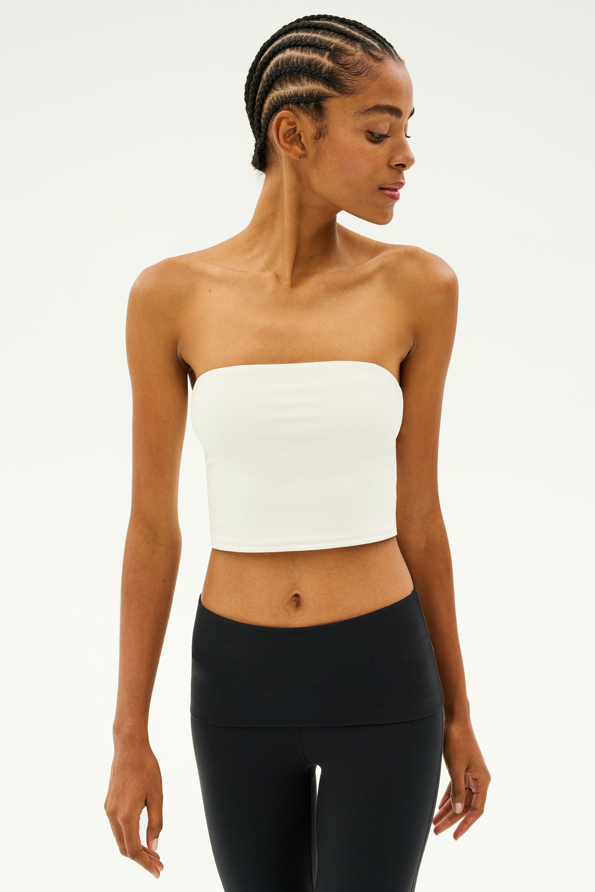 A person with braided hair wearing the SPLITS59 Hazel Airweight Crop Bandeau in white and black leggings, embodying flexibility as they look to the side against a plain background.