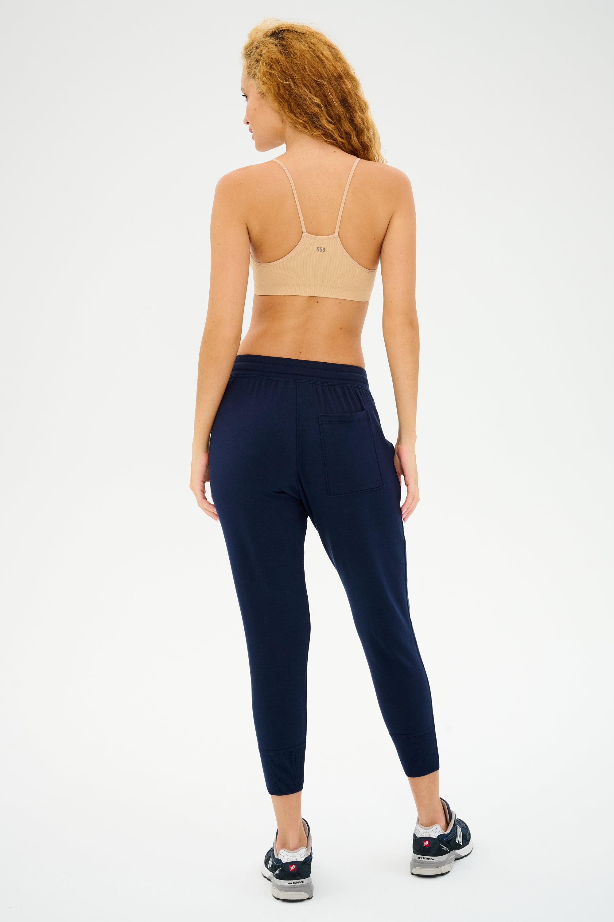 A person with curly hair is wearing everyday wear: a beige sports bra and the Splits59 Reena Fleece 7/8 Sweatpants in Indigo, standing against a plain background.