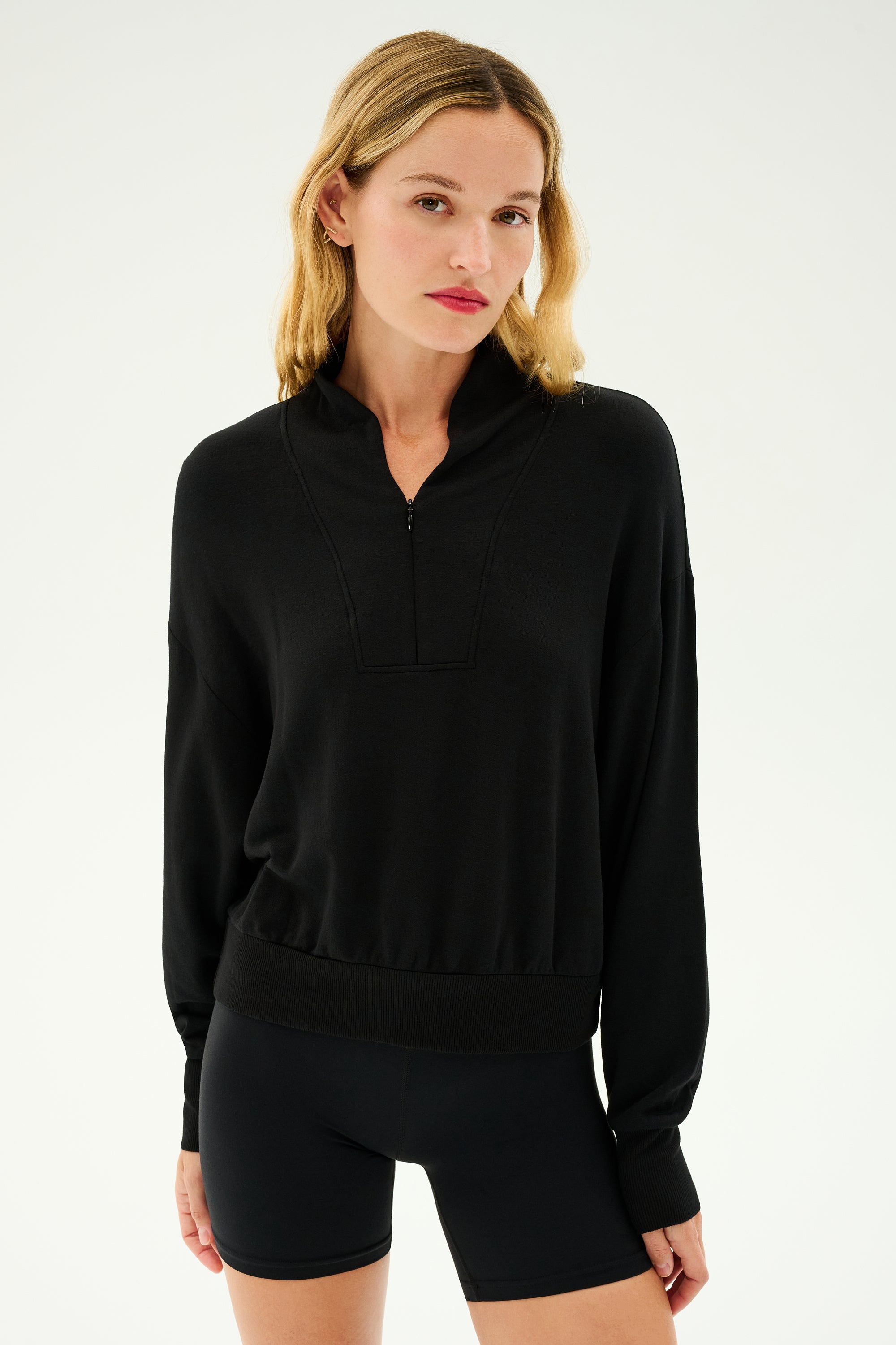 A person with shoulder-length blonde hair wears the SPLITS59 Birdie Fleece Half Zip in black, paired with black shorts, standing against a plain light background.