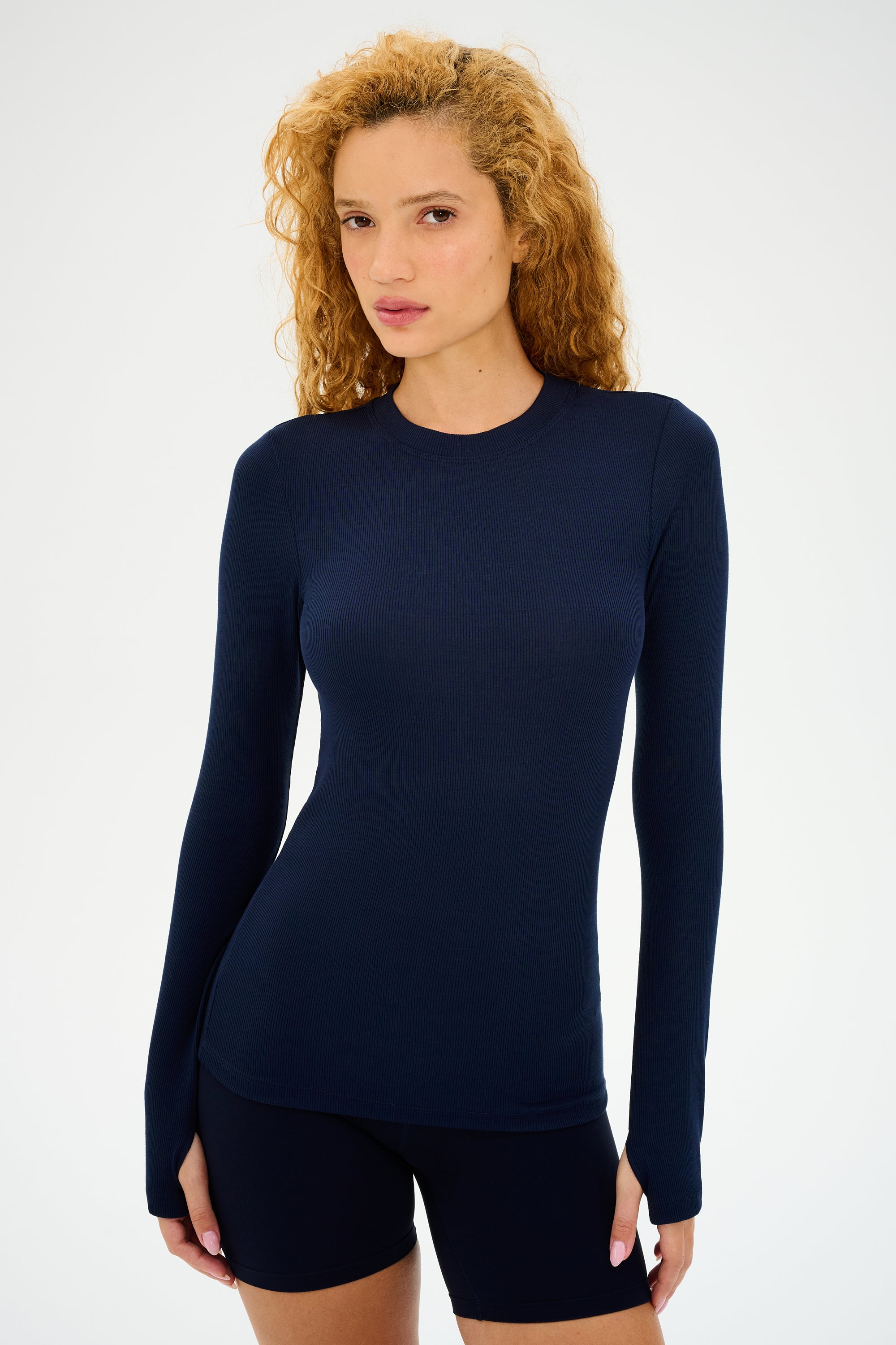 A curly-haired person stands against a plain background wearing the super soft Louise Rib Long Sleeve in Indigo from SPLITS59 and black shorts.