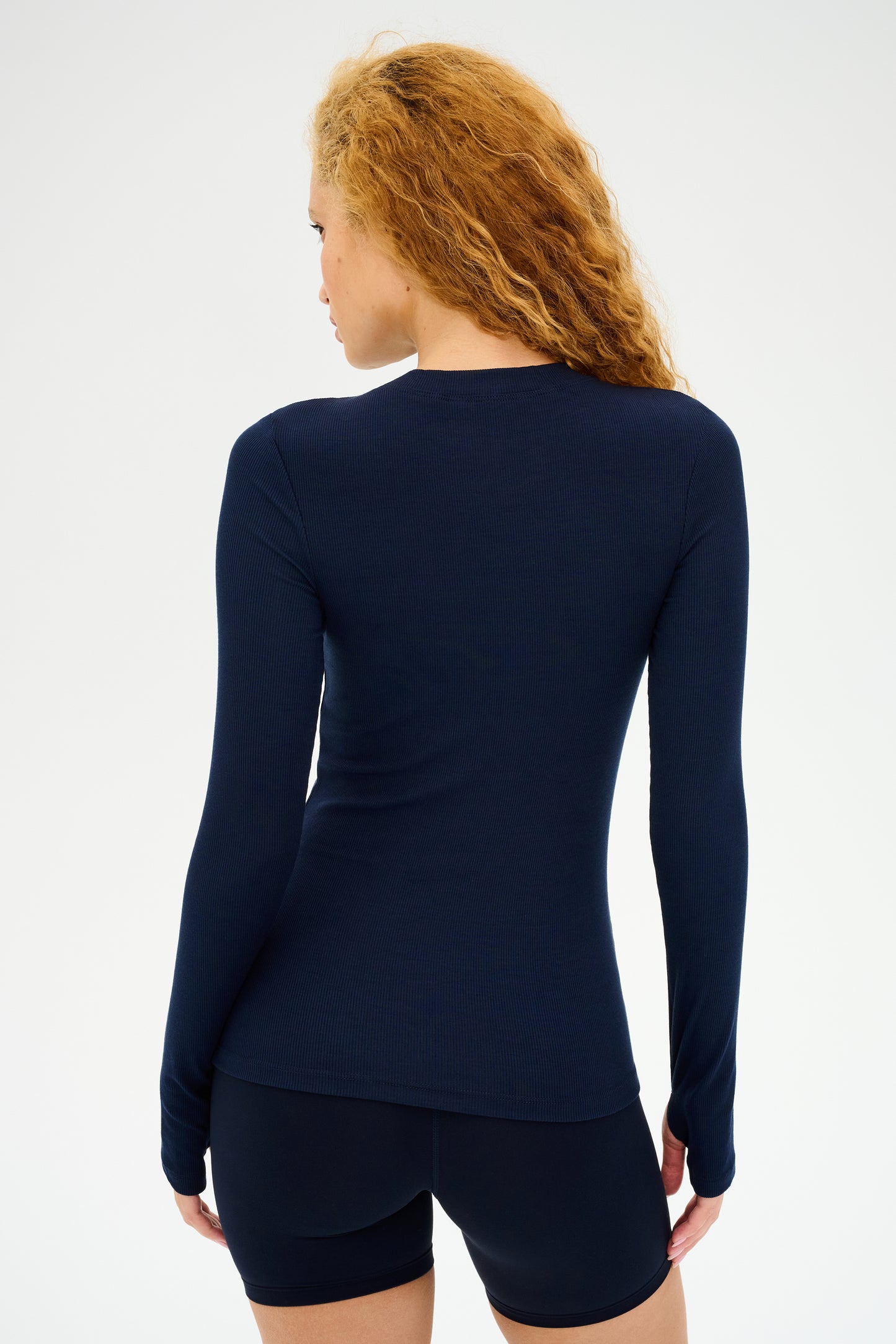 A person with curly hair stands facing away, wearing the SPLITS59 Louise Rib Long Sleeve in Indigo and black shorts on a plain background.