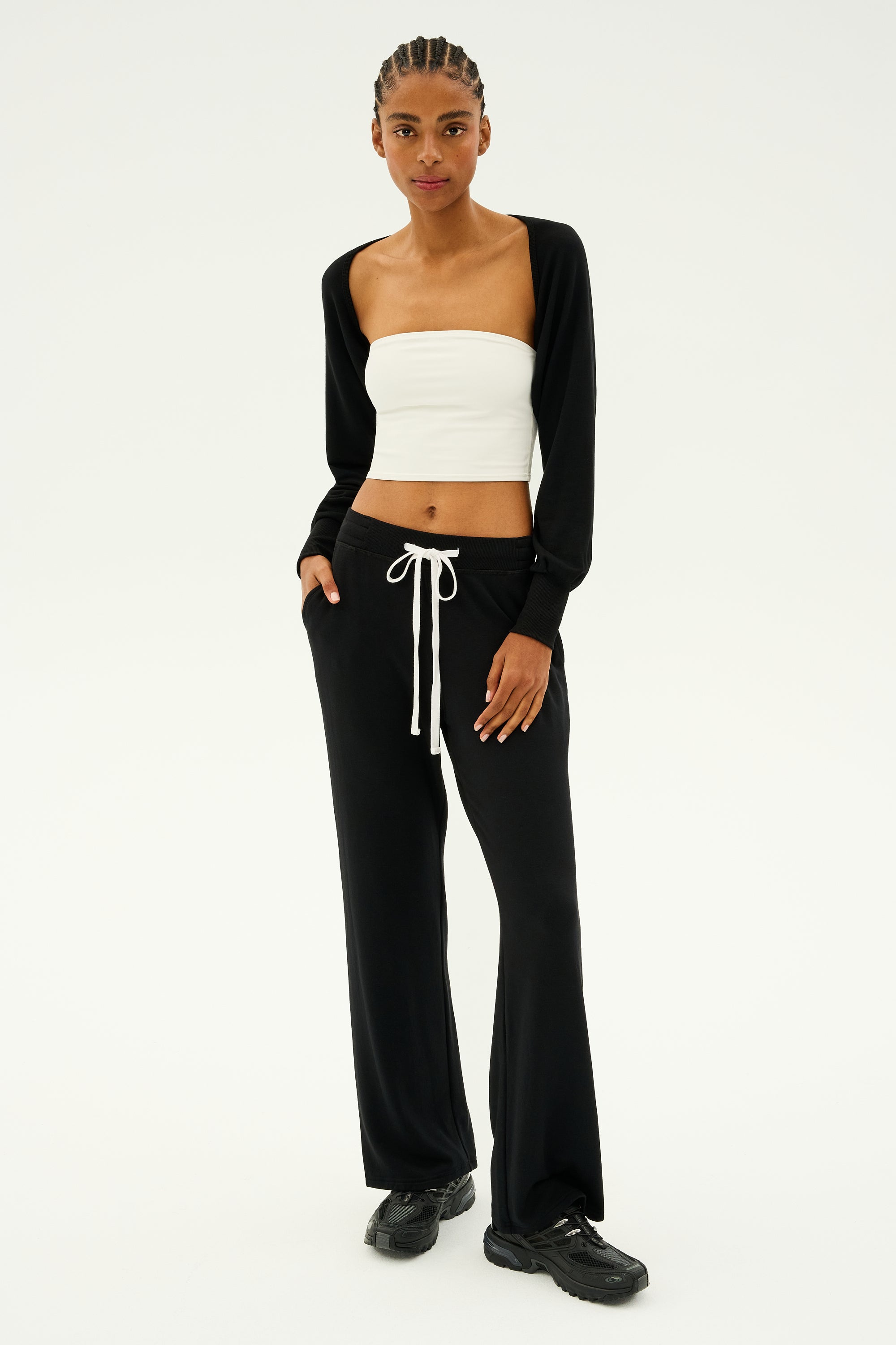 A person poses gracefully against a plain background, as if ready for yoga pilates barre, wearing a white tube top, black pants, and the SPLITS59 Sue Fleece Shrug in black.