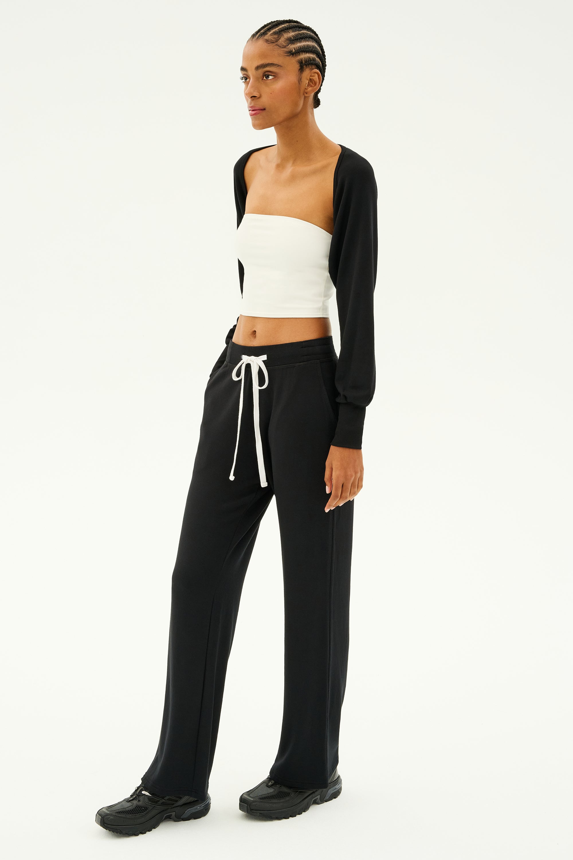 A person wearing the SPLITS59 Sue Fleece Shrug in black, paired with a white tube top and black drawstring pants ideal for yoga, Pilates, or barre classes, stands against a plain background in black shoes.