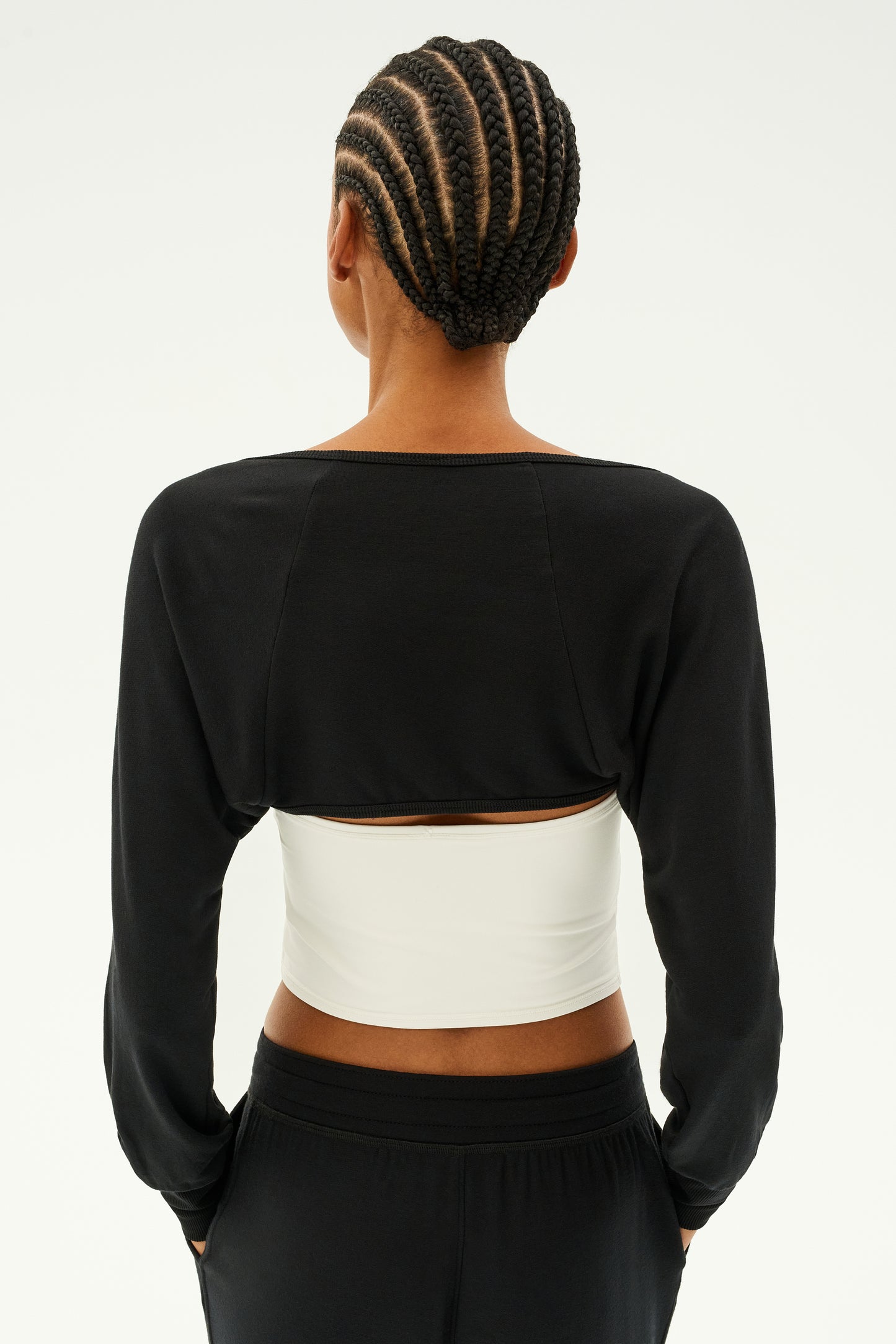 A person with braided hair, seen from behind, is wearing the SPLITS59 Sue Fleece Shrug in black over a white underlayer as they prepare for yoga pilates barre against a light background.