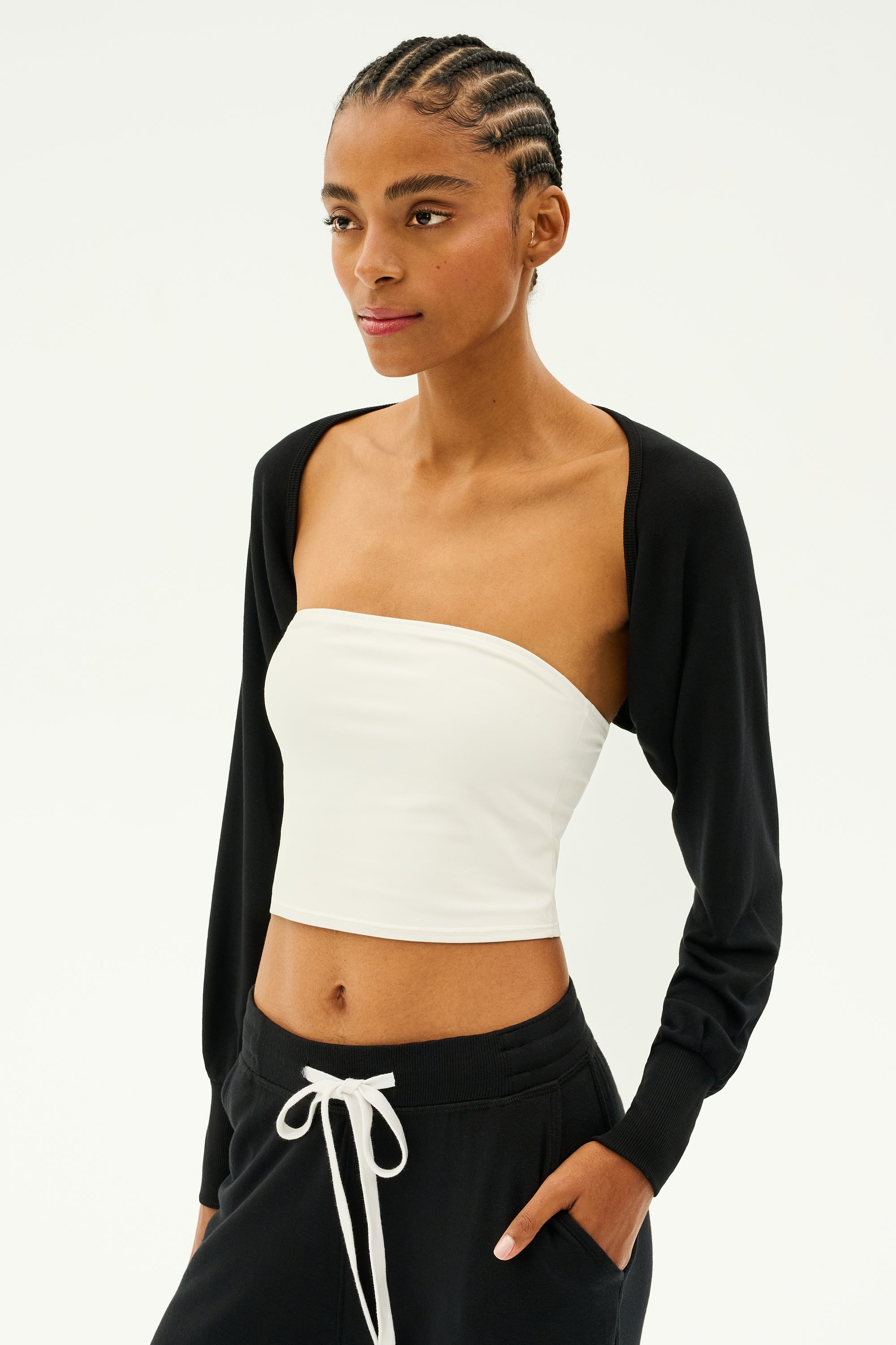 A person with braided hair is styled in a white tube top and black sleeves, combined with black pants featuring a white drawstring, ideal for dance warmups. The look is elevated by the chic Sue Fleece Shrug in Black from SPLITS59, emphasizing a sophisticated style made in Los Angeles.