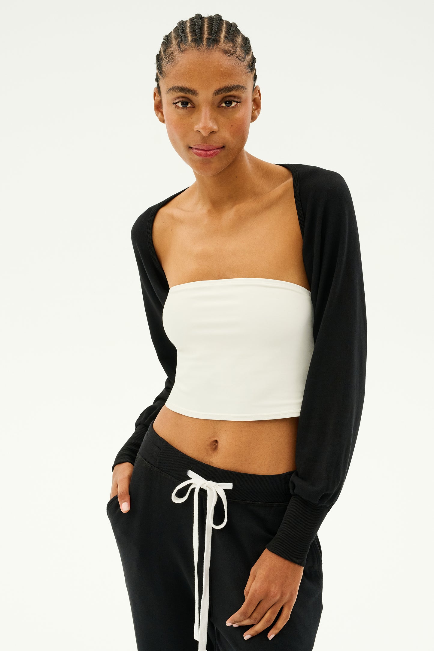 Dressed in the Sue Fleece Shrug - Black by SPLITS59 over a white strapless top and black pants with a white drawstring, the person stands gracefully against a plain background, exuding an aura of elegance as if ready to flow into a dance.