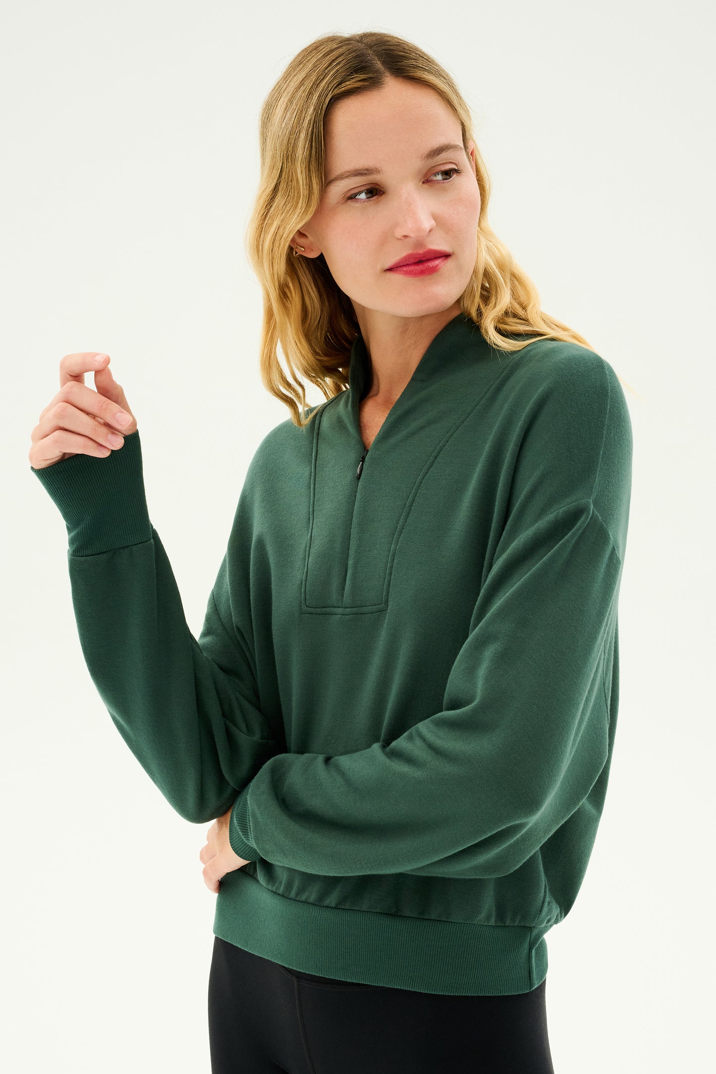 A woman with wavy blonde hair is wearing the Birdie Fleece Half Zip - Military by SPLITS59 and black pants. She looks to her left while gently holding the cuff of her sleeve with her right hand against a plain background.