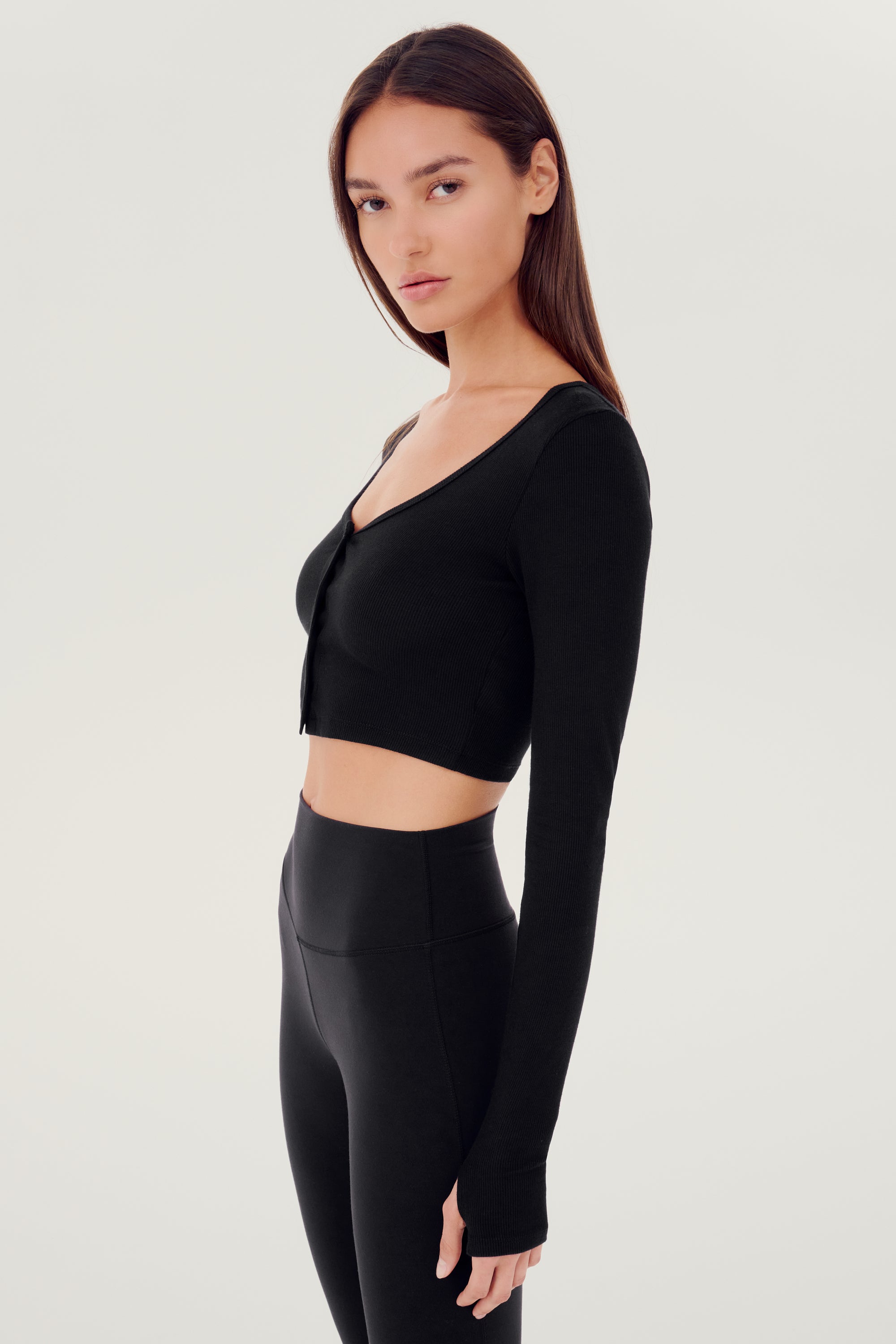 Front side view of woman with dark straight hair wearing cropped black four buttoned long sleeve cardigan with thumbholes and black wide bottom leggings.