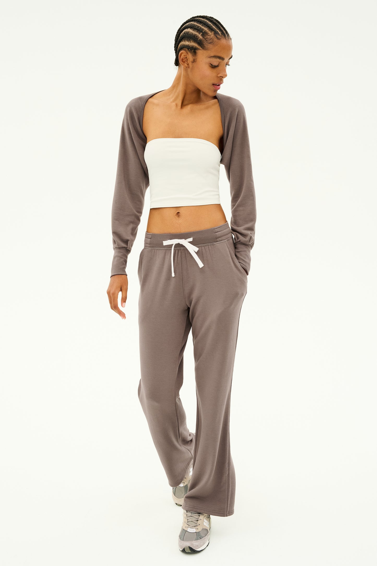 A person dressed in a white tube top, brown cardigan, and matching brown pants with sneakers stands against a plain background. This outfit is ideal for dance warmups or layering under the SPLITS59 Sue Fleece Shrug in Lentil.